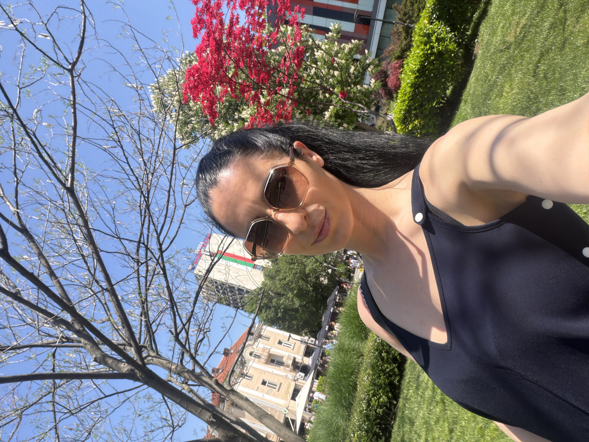A woman is taking a selfie in the city of Pleven, Bulgaria. She is standing on a grassy area. A tree with bare branches is to the left. A building with a Bulgarian flag on top is in the background, as well as other buildings. Red flowering trees are also in the background. The woman is wearing sunglasses and a dark-colored outfit with white buttons.