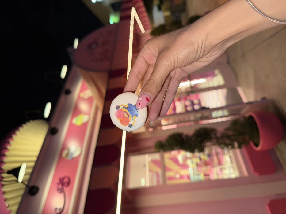 A human left hand is positioned centrally, holding a circular white macaron. The macaron features an edible print of a yellow Minion character, dressed in blue overalls, balancing two scoops of ice cream (one pink, one orange) on its head. The hand's fingernails are manicured with a light polish base and small red heart decals are visible on the thumb and index finger. A thin, silver-colored bracelet is worn on the wrist. The background, out of focus, depicts the exterior of a brightly lit dessert shop under a dark night sky. The building is predominantly pink, featuring a striped awning designed to resemble a large cupcake liner, beneath which are illustrative depictions of various desserts. Partial branding for "Hello Kitty" is visible on the right side of the awning. The shop's entrance consists of glass doors revealing a vibrant, well-lit interior with pink and white striped elements and shelves stocked with confectionery. A potted, spiral-shaped topiary plant in a pink container is situated to the left of the entrance. This location is identified as Hello Kitty's Cafe in Universal Studios Florida, Orlando, United States of America.