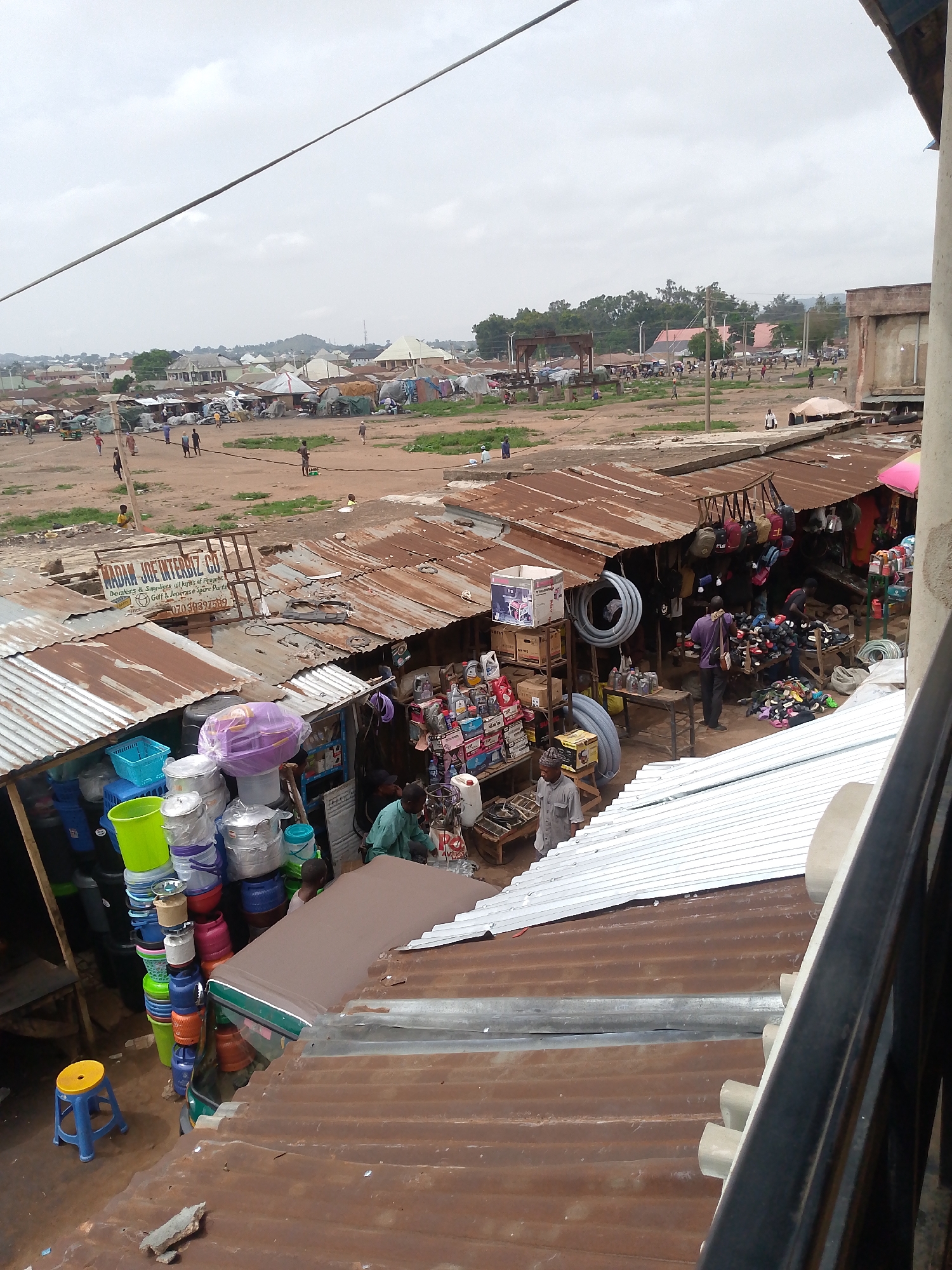 The image captures a bustling market scene in Bukuru, Nigeria, from an elevated vantage point. Below, rows of market stalls with corrugated iron roofs are packed tightly together. One stall displays a colorful array of plastic buckets and basins. Another stall, identified by a sign as "MADAM JOF INTERBIZ CO," deals in "Peupebt, Golf & Joperase Spare Parts." A man in a purple shirt browses through a shoe stall, while another man in a grey shirt stands near a stall selling engine oil. In the background, a large dirt area is visible, with scattered buildings and people in the distance. The sky overhead is overcast.