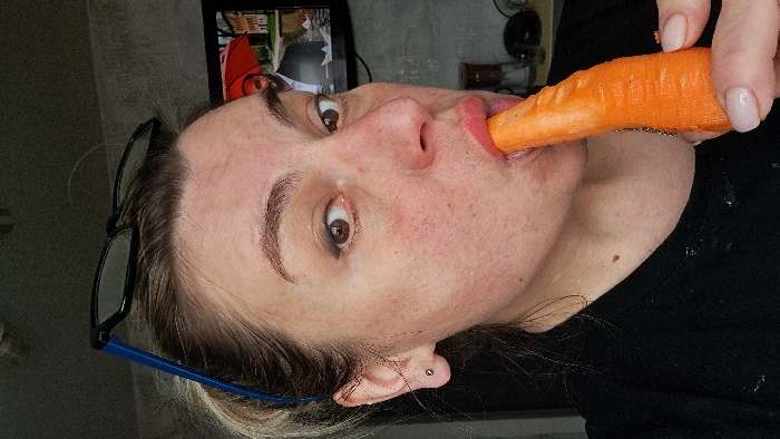 An adult female is depicted in a close-up, chest-up shot. She has light brown hair pulled back, dark brown eyes with visible eyeliner, and some facial redness or freckling. Blue-templed, black-framed glasses rest on top of her head. A small gold stud earring is present in her left ear. She wears a black garment. Her right hand, with light pink nail polish, holds an orange, peeled carrot, the thicker end of which is placed into her pursed lips. In the blurry background, a television screen displays a male figure in a suit standing under a red umbrella, with small white objects resembling paper boats nearby. Indistinguishable household objects, possibly containers, are visible on a surface beneath the television. The wall surface is a light, textured grey. The specific location within the city of Bel'kovo, Russia, is not discernible from the image.
