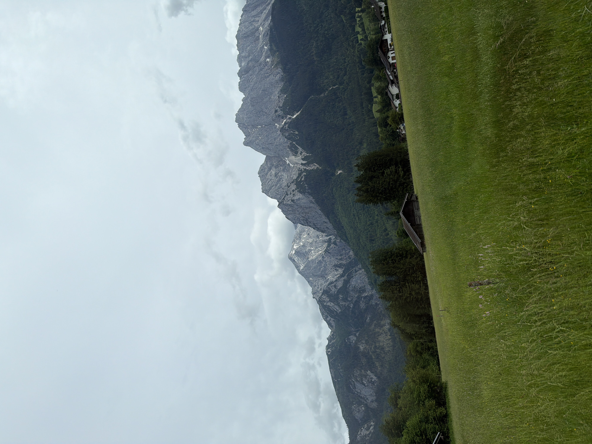 The image, oriented incorrectly and appearing rotated 90 degrees counter-clockwise, depicts a vast, serene mountainous landscape under an overcast sky. When viewed correctly in a horizontal landscape orientation, the scene features a large, lush green field in the foreground, extending from the right side of the frame towards the left. The grass appears tall and thick, with hints of lighter stalks and small yellow wildflowers. In the lower right corner of the field, a small, rustic wooden structure, possibly a shed or shelter, is partially visible behind some dark green trees.
Beyond the field, a majestic range of mountains dominates the midground and background. These mountains are rugged and rocky, with grey and dark grey tones, and some patches of snow or light rock are visible on their upper slopes. Dense, dark green forests cover the lower elevations and valleys of the mountains. On the right side of the image, nestled at the base of the mountains or on a gentle slope, is a cluster of buildings, likely houses or chalets, characterized by white walls and brown roofs, indicating a small village or settlement.
The sky above the mountains is heavily overcast with light grey clouds, suggesting a cloudy day. There are no harsh shadows, consistent with an overcast sky. No people or specific activities are visible, emphasizing the natural, tranquil beauty of the landscape. There is no visible text in the image. The architectural style of the houses and the mountainous terrain are consistent with the provided location context of Grainau, Germany.