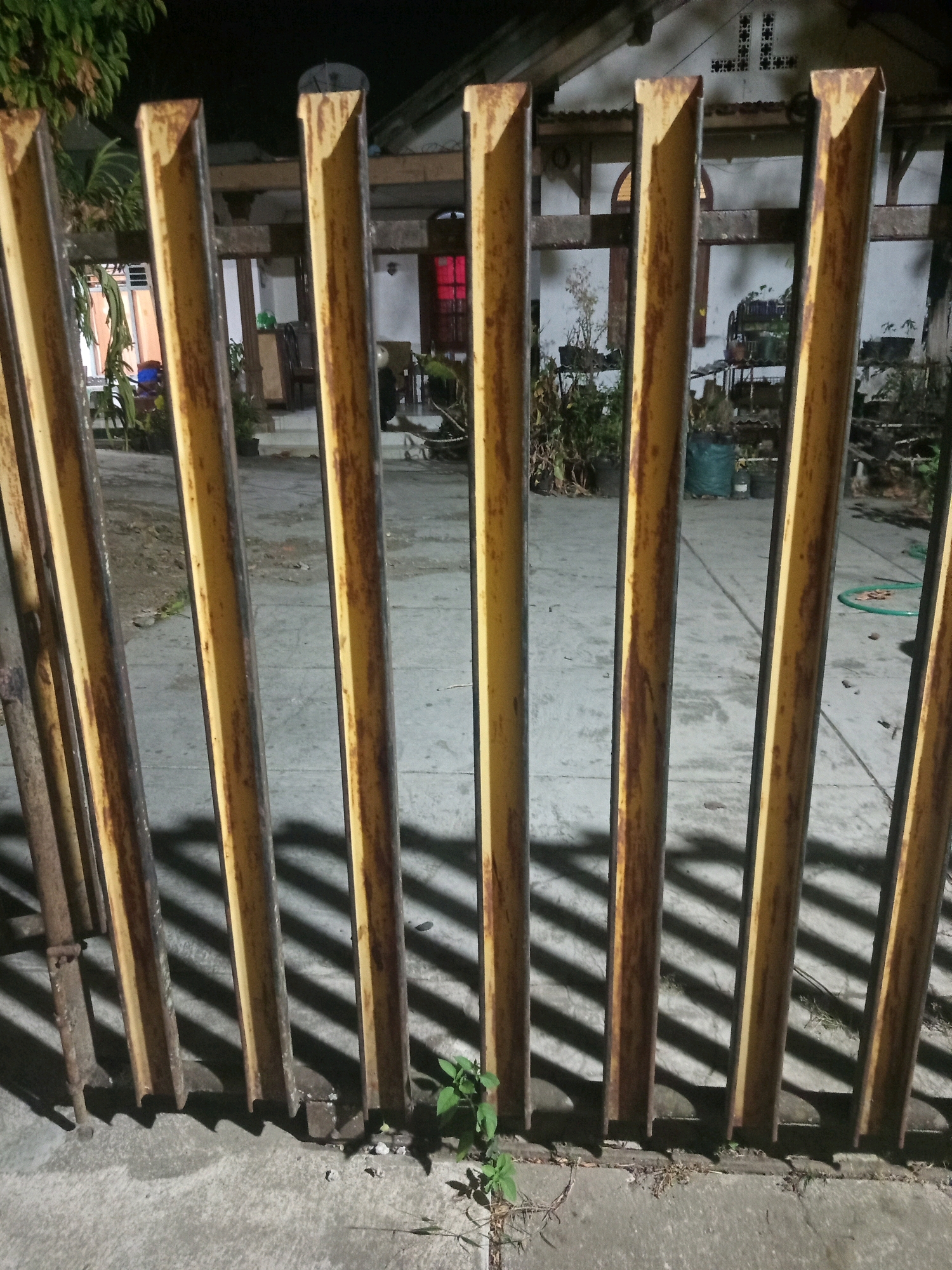 A perspective shot taken at night shows a rusty, yellow metal fence running diagonally across the foreground. The fence is composed of vertical I-beams, with shadows cast by a light source to the left, creating a striped pattern on the concrete ground behind the fence. In the background, through the fence, a house with an open doorway revealing a red light can be seen. Plants and some potted vegetation are visible in the yard. The scene depicts the exterior of a property in Ketindan, Indonesia.