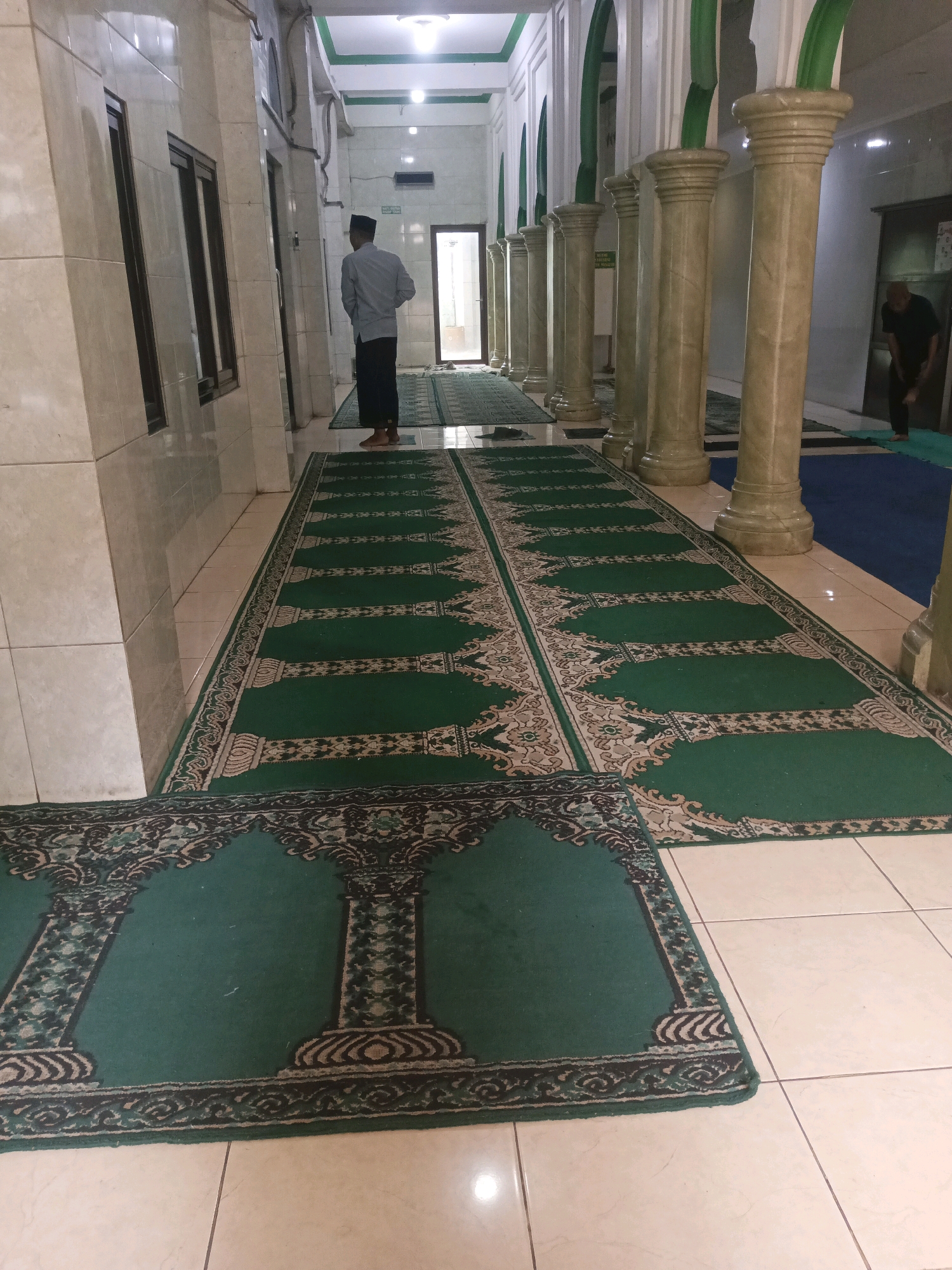 The image depicts the interior of a mosque, possibly located in South Tangerang, Indonesia, with a focus on prayer rugs and architectural elements. In the foreground, a green prayer rug with a black and white intricate border and a pattern resembling arches and columns is laid out on a tiled floor. Behind it, a longer, similarly patterned green prayer rug stretches down a hallway, with alternating dark green and lighter green designs forming prayer niches.
Several ornate, beige-colored columns with decorative capitals line the right side of the hallway, supporting an arched ceiling structure which is painted white with green accents. The hallway walls are a light beige tile.
Two individuals are visible in the scene. On the left, a person wearing a light blue long-sleeved shirt and dark pants, with a black skullcap, stands with their back to the camera, facing towards the end of the hallway. On the right, further down the hallway and partially obscured by columns, another person in dark clothing is bending over, possibly preparing to pray or adjust a rug. The lighting appears to be artificial, casting a slight yellow hue on the scene. The environment suggests a place of worship, with the emphasis on the prepared prayer spaces.