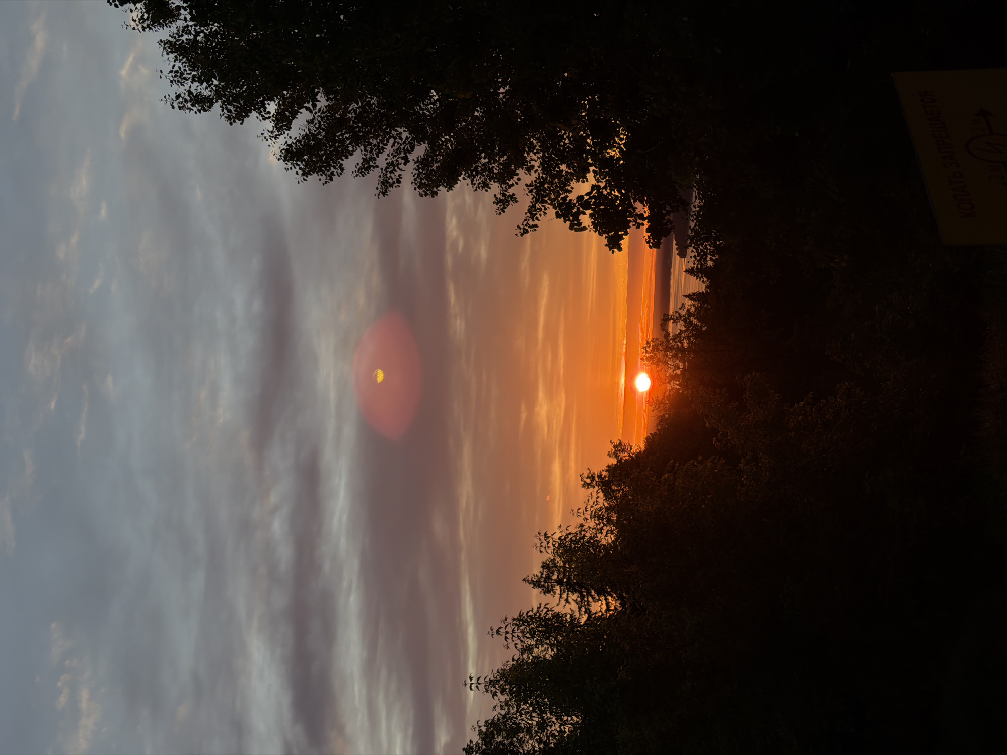 The image depicts a sunset viewed through trees, possibly in Izhevsk, Russia, given the Russian text on a sign in the right corner. The sun appears as a bright orange orb on the horizon. The sky is a mix of gray clouds and orange hues, transitioning from darker to lighter towards the horizon. A lens flare appears above the sun. The surrounding trees are dark silhouettes framing the sunset.