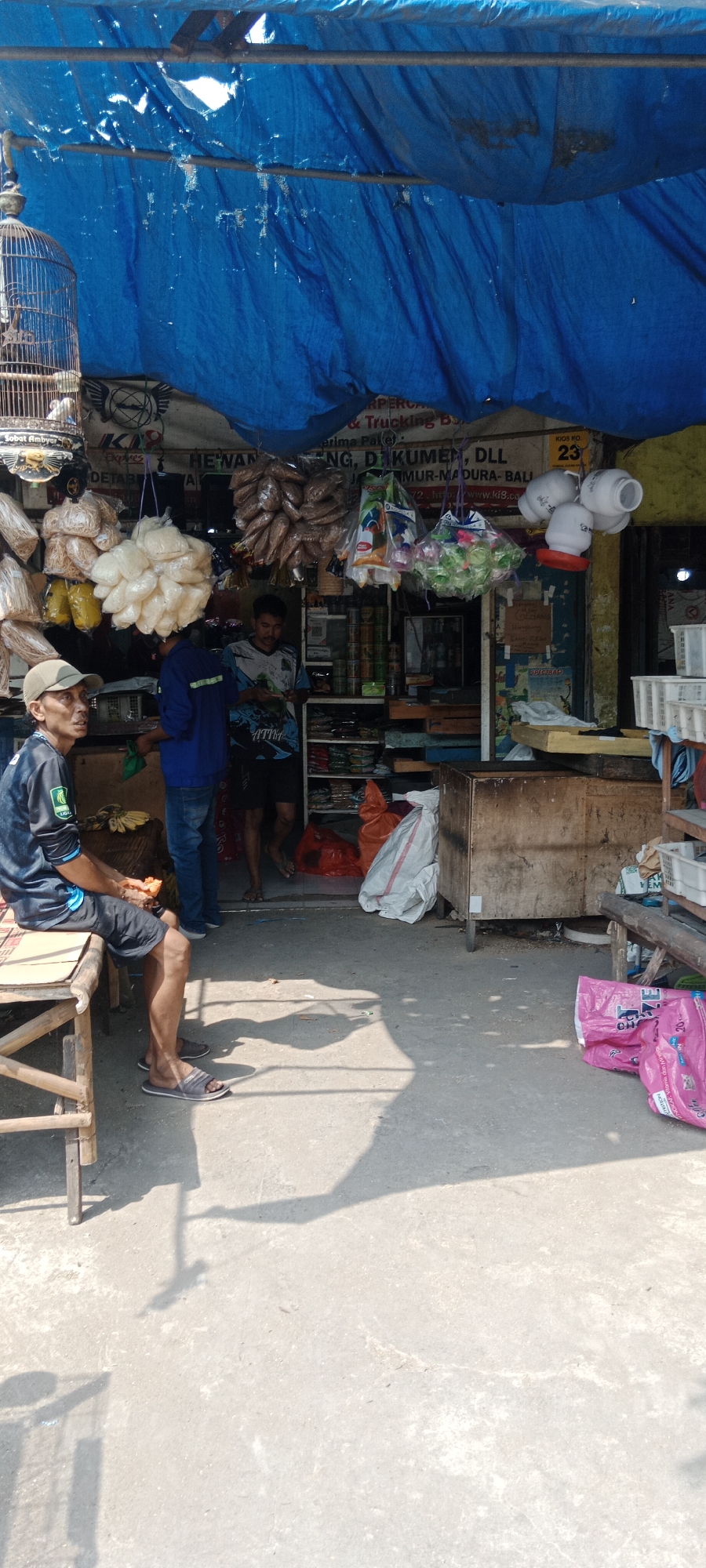 The image shows an outdoor storefront in Tumenggungan, Indonesia, likely a small market or shop selling various goods. A bright blue tarp, partially ripped and faded, is stretched overhead as a makeshift roof, supported by wooden poles. The store's interior is visible, filled with shelves stocked with packaged goods. In front of the store, there are several hanging items, including a birdcage with a bird inside, bags of what appear to be dried goods or snacks, and plastic bags containing colorful items.
A man wearing a dark long-sleeved shirt with green accents and a light brown cap sits on a wooden bench to the left of the frame, facing away from the camera. He is wearing dark shorts and grey sandals and is holding what looks like a piece of food or an object in his right hand. He is casting a shadow on the concrete ground.
Inside the store, two other men are visible. One man in a blue long-sleeved shirt and jeans is standing near the entrance. Another man, wearing a patterned t-shirt and dark shorts, is standing further inside, looking down at something in his hands.
Various objects are scattered around the storefront, including large sacks, plastic bags (one bright pink bag with text is prominent on the right side of the frame), wooden crates, and what appear to be animal feeders hanging from the tarp. The ground in front of the store is concrete and shows significant shadows from the overhead structures and people. The overall scene suggests a busy, everyday commercial setting.