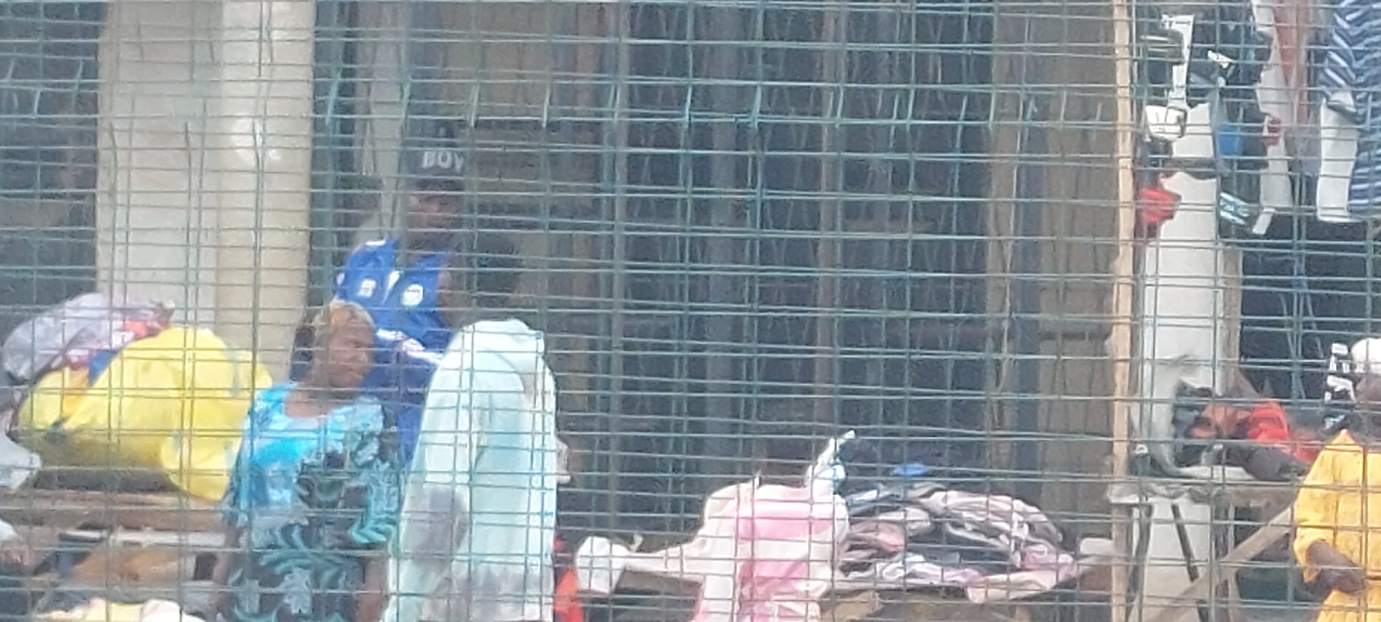 The image captures an outdoor market or street vendor scene through a prominent wire mesh fence, located in the city of Unknown, Unknown. In the left-center, a person wearing a blue collared shirt and a dark cap with the word "BOY" printed on it stands. To their left, a woman with light-colored hair, wearing a blue and black patterned dress or top, is visible. A large, light-colored fabric or garment is draped vertically in the central foreground. Numerous piles of assorted clothing and textiles in various colors, including yellow, pink, gray, red, and blue with striped patterns, are displayed on makeshift surfaces across the scene, particularly to the right. An individual partially clad in a yellow garment is visible on the far right. Indistinct building structures are visible in the background, suggesting an urban environment.
