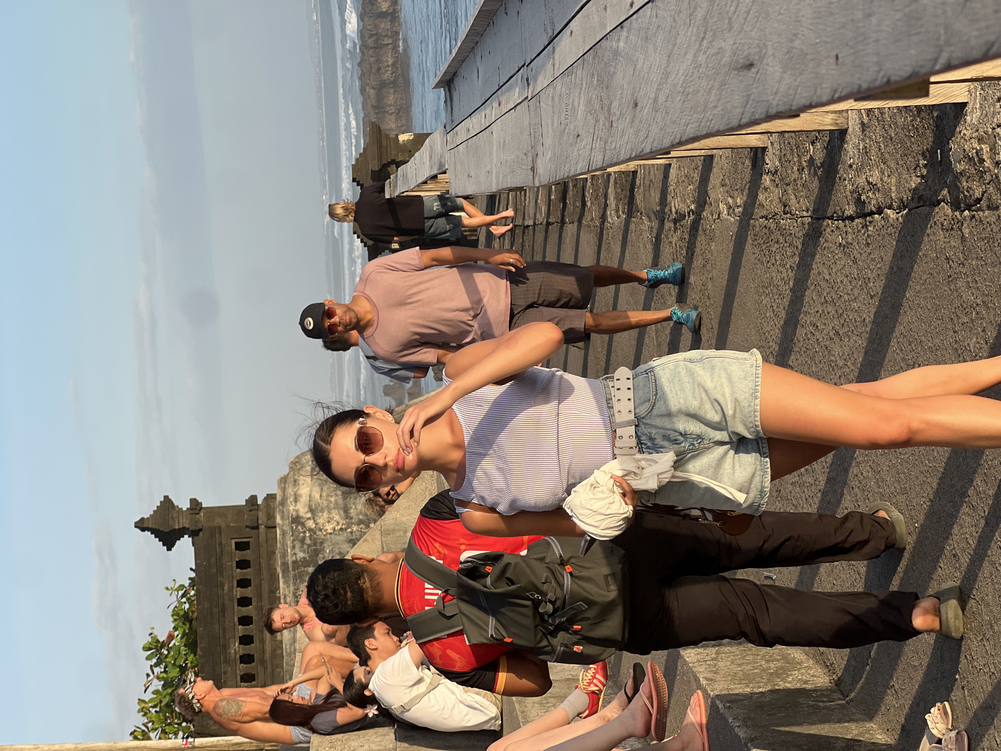 In North Kuta, Indonesia, a group of people are seen near a stone structure with ornate carvings, possibly a temple, overlooking the ocean. The sky is a soft blue with wispy clouds. The foreground features a woman in the center wearing a striped top and denim shorts, holding a white cloth. She has on sunglasses and a belt. To her left, a man in a red shirt and black pants with a backpack walks away from the camera. To the left of the man, several people are seated on stone steps. In the background, a man in a pink t-shirt and shorts walks towards the ocean, and further in the distance, another person is seen on a wooden boardwalk. The ocean has waves, and the lighting suggests it is late afternoon or early evening.