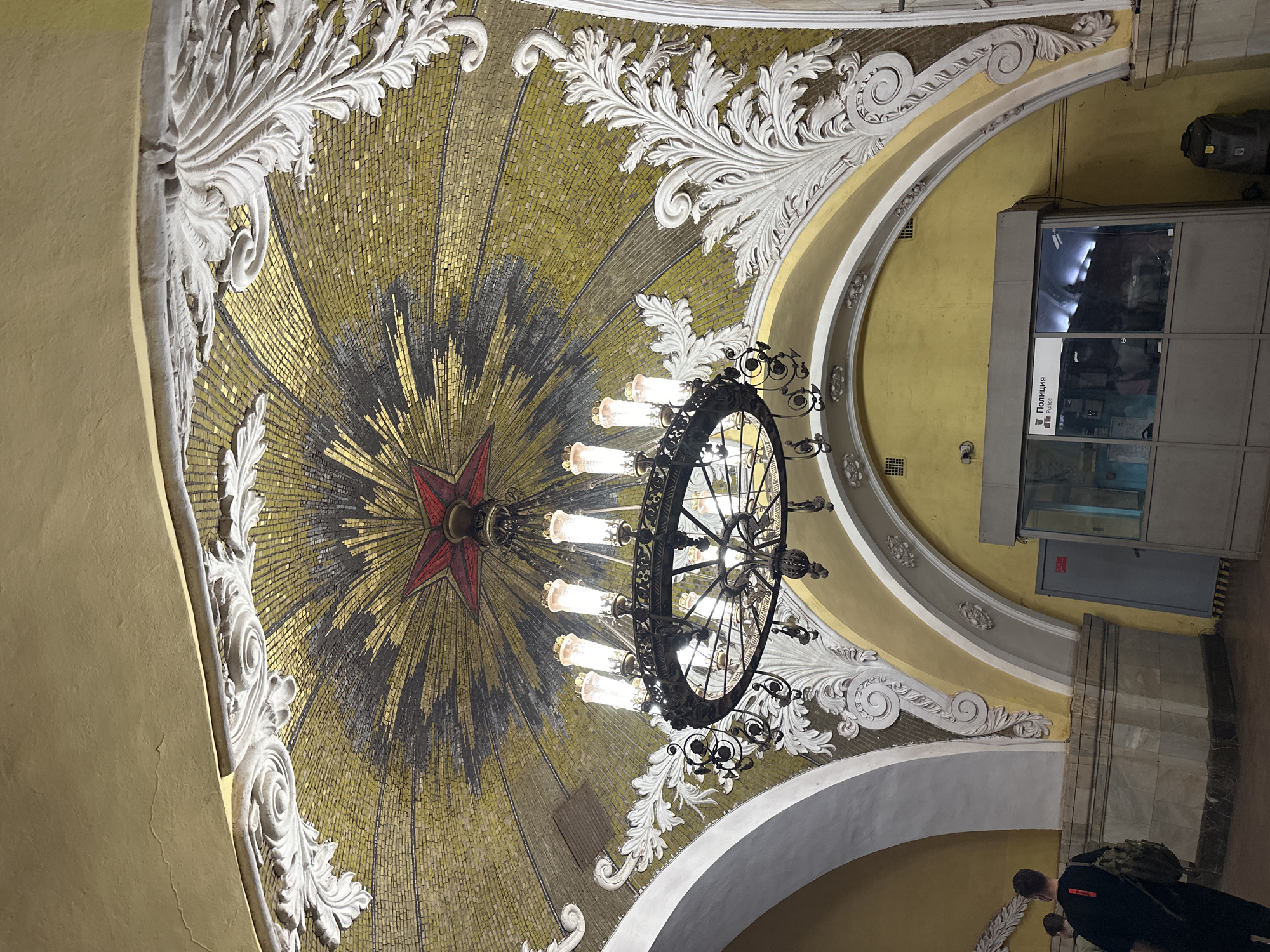 The image shows the ceiling of a Moscow metro station in Russia. A large, ornate chandelier hangs from the center of a mosaic dome. The dome is primarily composed of gold and yellow tiles, forming a starburst pattern with a red star at the center. Decorative white plasterwork surrounds the dome. To the right, a Police box. On the bottom right, a man walking away wearing dark clothing and a backpack can be partially seen. The image is taken from a slightly low angle, looking upwards.