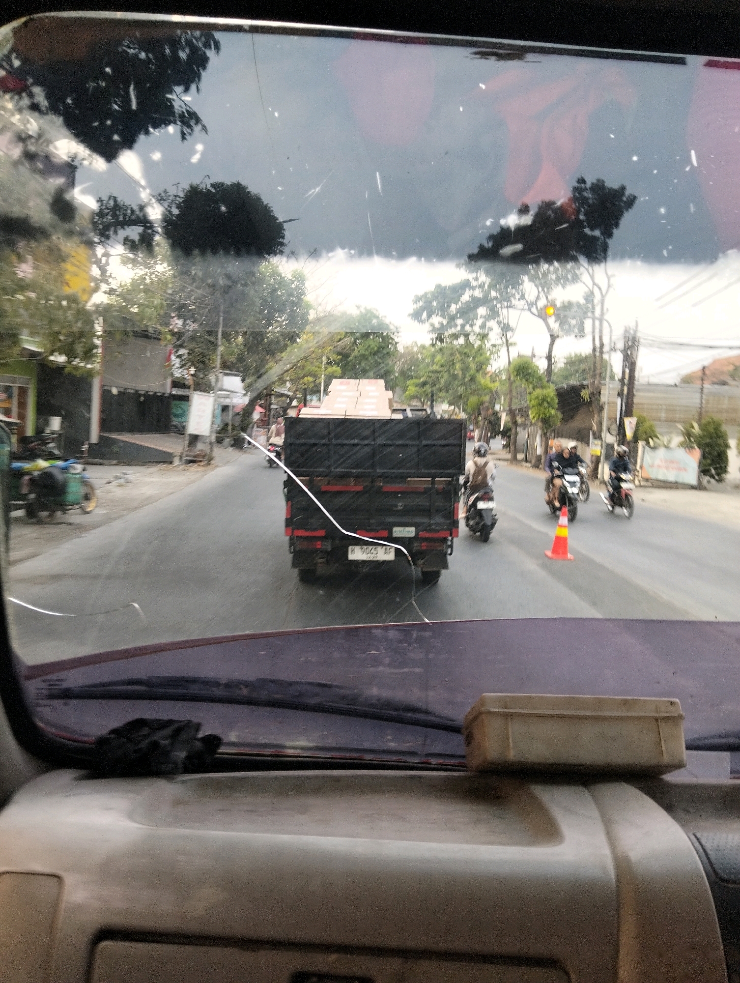 The image is a rear-view perspective from inside a vehicle, looking through the windshield. A dark-colored truck with a black cargo box is in the foreground, traveling on a paved road. The truck has a license plate that appears to read "H 9045 AF". In the middle ground, several motorcycles are also on the road, with riders visible on each. One motorcycle in the center has an orange traffic cone positioned behind it. The road is lined with buildings on the left side and trees and utility poles on both sides. The sky is overcast. On the dashboard of the vehicle in the foreground, a small, light-colored rectangular box is resting. The windshield has several visible cracks and smudges. The scene is consistent with a street in Semarang, Indonesia, as indicated by the license plate format and the general urban and tropical environment.