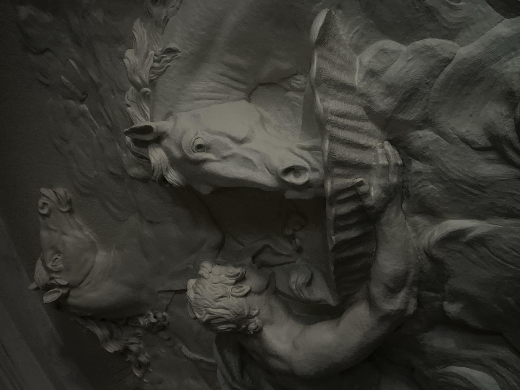 The image depicts a bas-relief sculpture featuring two rearing horses and a partially nude male figure. The scene is rendered in a monochrome gray tone, mimicking the appearance of stone or plaster. The male figure is positioned below one of the horses, seemingly restraining it with one arm while holding a ridged, board-like object in the other hand. The horses are depicted with detailed manes and expressive features. The overall composition suggests a dynamic and dramatic interaction between the human figure and the animals. This bas-relief is located in Saint Petersburg, Russia.