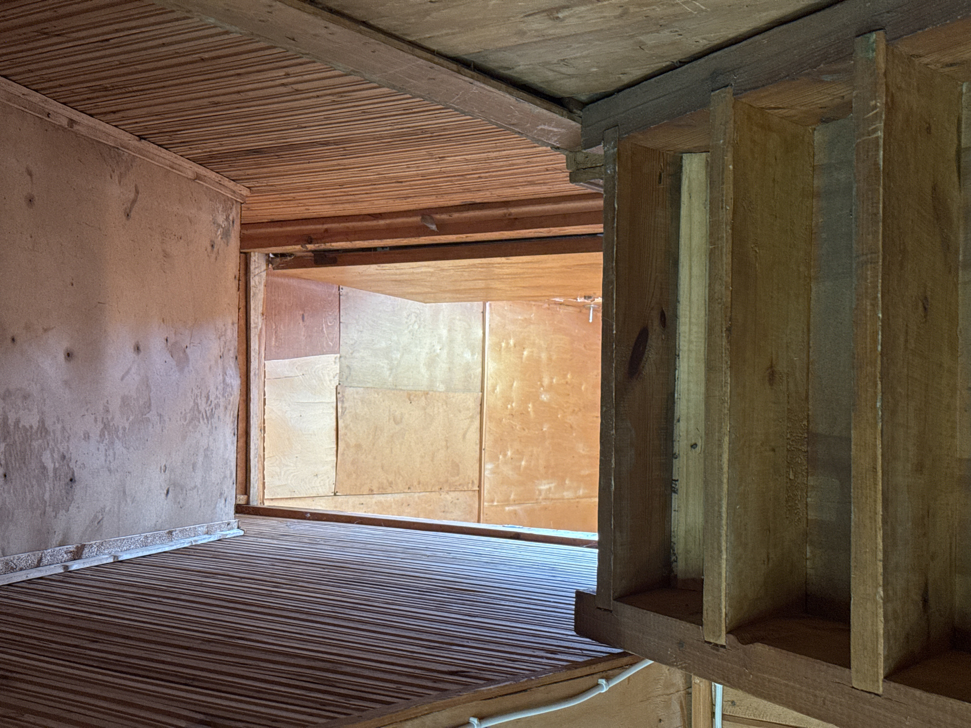 The image shows the interior of a structure, possibly a room or a compartment, built primarily of wood. The floor is made of narrow wooden planks or slats, laid in parallel, with dark stripes running along their length. The ceiling is also made of wooden planks, laid in the same direction as the floor. The walls are constructed of large plywood sheets, showing variations in color and texture.
In the foreground, a wooden frame or structure with vertical planks creating compartments is visible. The lighting is uneven, with some areas appearing brighter than others. The room seems aged and possibly neglected. There are no people or clear subjects present in the image. The location is marked as Unknown, Unknown.