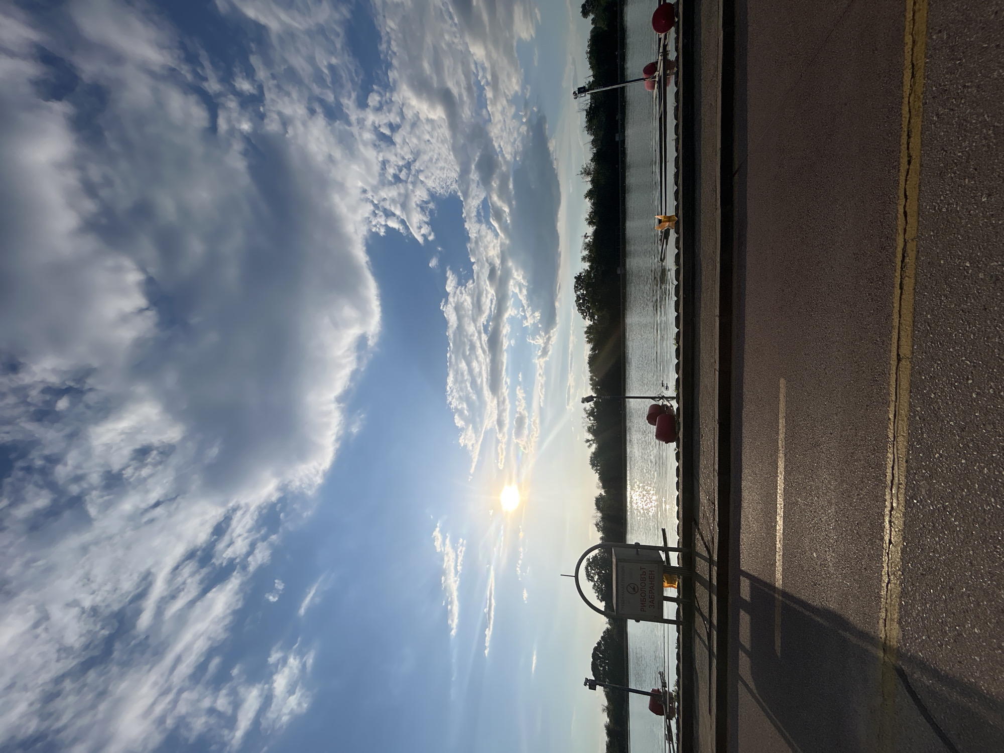 Here's a description of the image:
The image depicts a view of a body of water (likely the Maritsa River) with a partially cloudy sky above. A section of what appears to be a paved road or pathway is visible on the right side, marked with a yellow line. To the left of the road/pathway is a metal railing or barrier, separating it from the river. A sign with Bulgarian text is attached to the railing, which indicates “Fishing is prohibited.” In the distance, across the river, is a line of trees. The sun shines brightly, reflecting off the water. The location is likely along the Maritsa River in Plovdiv, Bulgaria, given the river's presence in the city and the Bulgarian text on the sign.