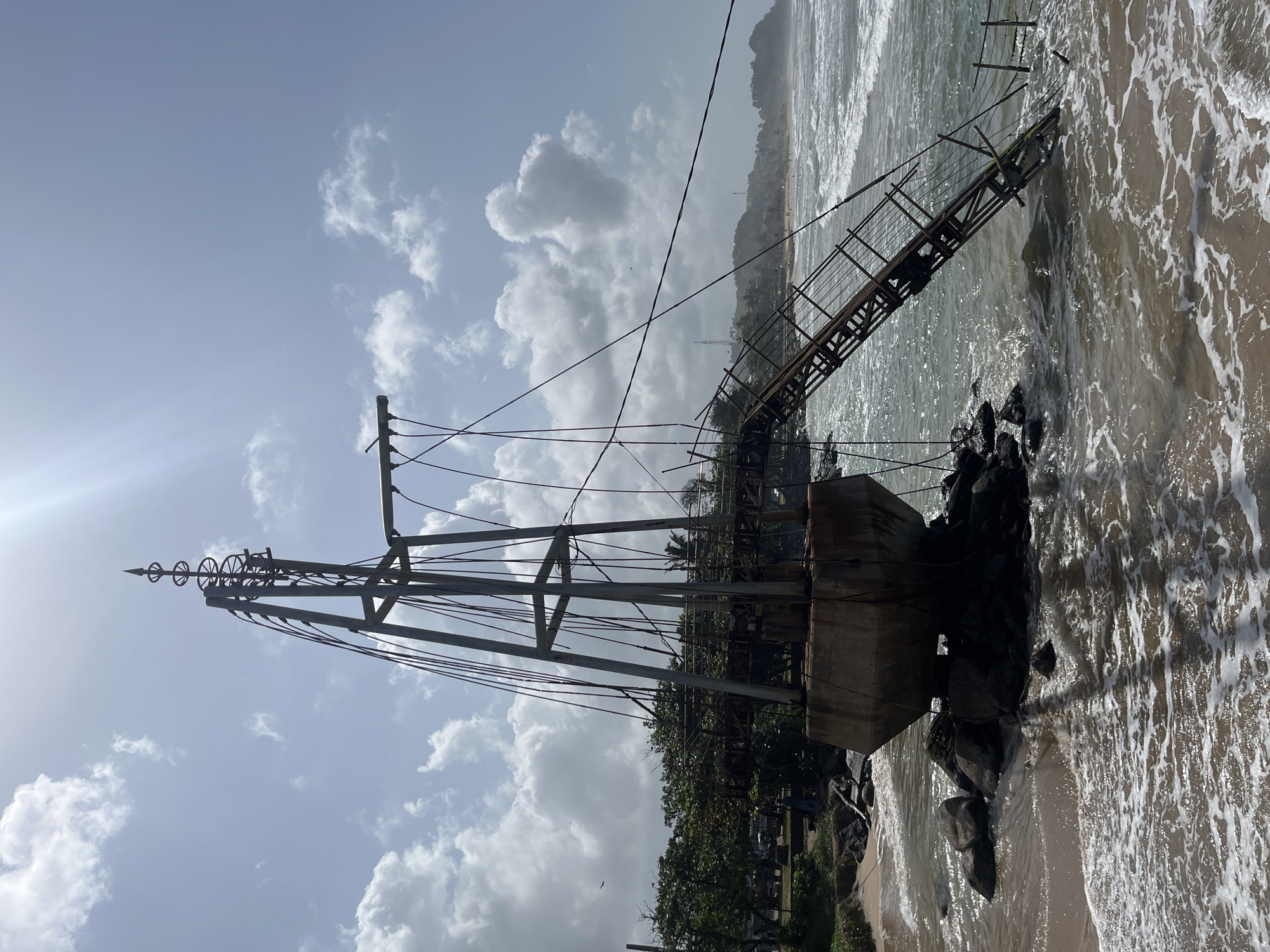 An aerial view shows a metallic structure, possibly a lighthouse or a navigational beacon, partially submerged in the ocean off the coast of Walgama, Sri Lanka. The structure, made of concrete and metal, is surrounded by rocks and appears weathered. A metal ramp extends from the main structure towards the water. The ocean is calm with gentle waves, and the sky is partly cloudy. In the background, a sandy beach is visible with trees and some buildings. The image appears to be taken on a sunny day.