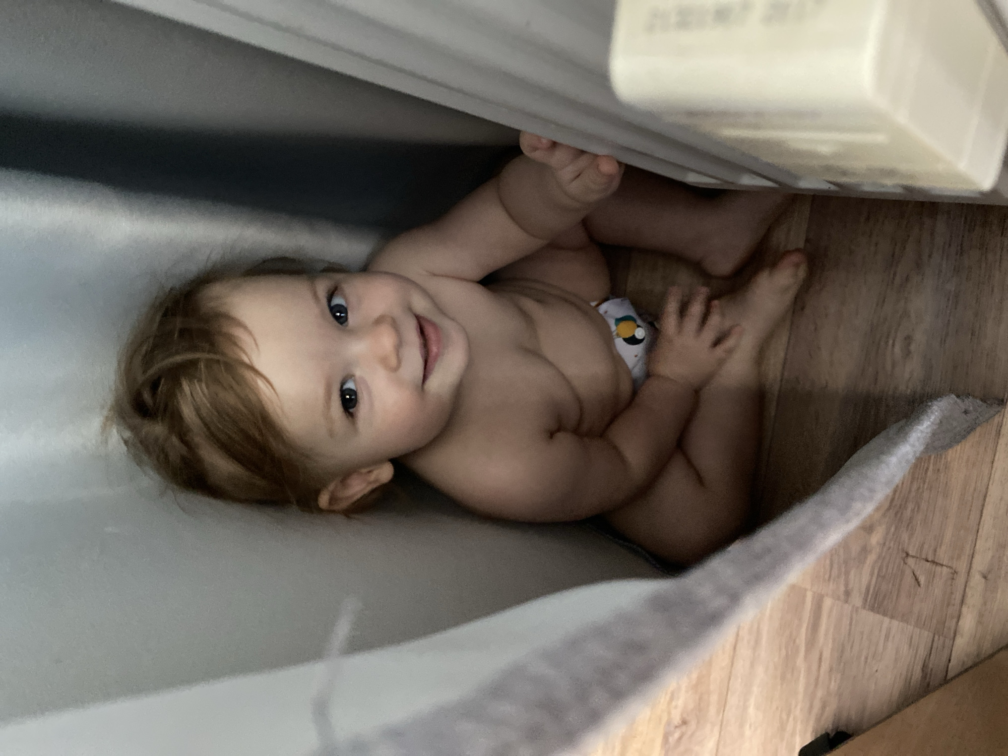 An overhead photograph captures a cheerful baby lying on their back in an indoor setting. The infant, with light brown hair and bright blue eyes, is looking directly at the camera with a wide, happy smile. They are partially clad, wearing a white diaper adorned with a small, colorful graphic, possibly a penguin.
The baby is situated on a soft, light gray fabric or blanket that covers most of the left side of the frame. On the right, a warm-toned wooden floor is visible beneath and around the baby's lower body. Above the baby's head, a large, light-colored object, possibly a piece of furniture like a crib rail or the side of a playpen, extends across the top of the image. This object creates a sense of the baby being nestled in a cozy, confined space.
Faint, light-colored text, which appears to be a date such as "24/07/2017" or a similar alphanumeric sequence, is visible on the surface of the object directly above the baby. The lighting in the scene is soft and ambient, illuminating the baby's face and features clearly. The overall mood is playful and endearing, capturing a moment of a content and curious child.