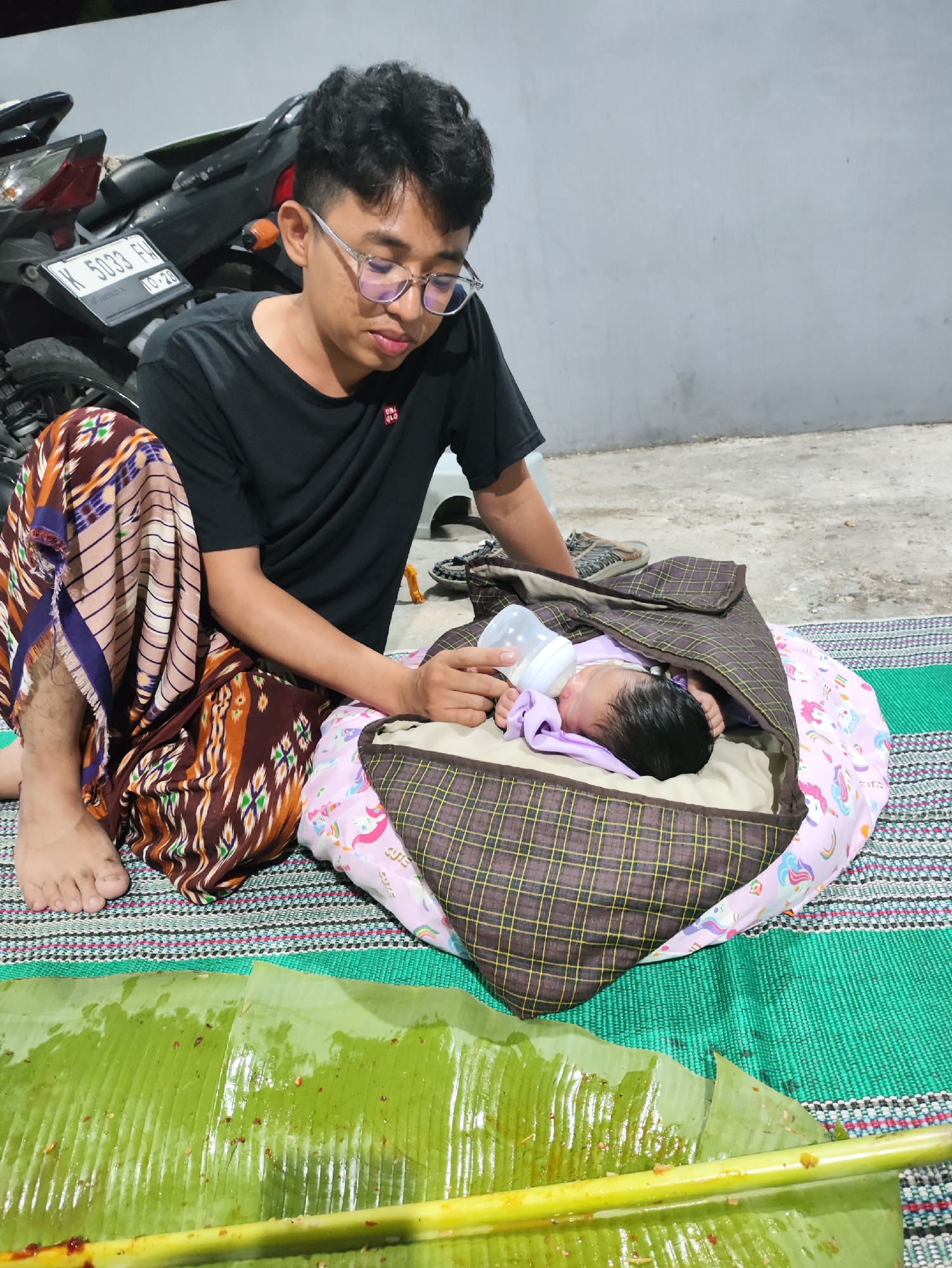 A young man wearing glasses and a black t-shirt is kneeling on a woven mat, feeding a baby from a bottle. The baby is nestled in a fabric carrier lined with a pink unicorn-patterned bib. Behind them, a motorcycle is parked against a grey concrete wall. In the foreground, a large banana leaf, glistening with moisture and speckled with food remnants, lies on the mat. The scene appears to be an outdoor setting, possibly a yard or patio, with a rough concrete floor. The overall impression is one of domestic care and simple living.