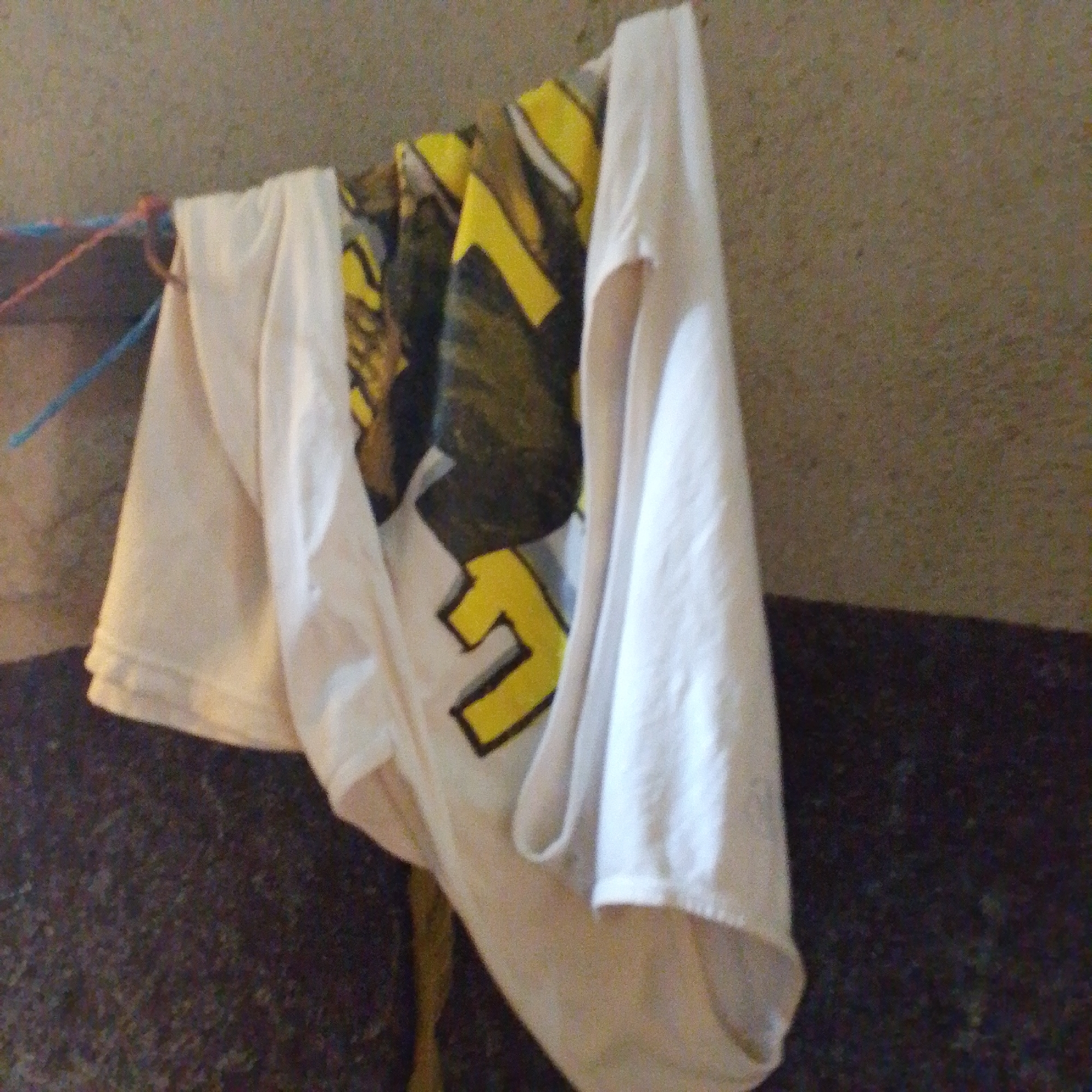 A white t-shirt with a yellow graphic is hanging on a clothesline. The t-shirt is partially obscured by another white garment. The background is a textured, light brown wall. The clothesline is made of blue and pink cord. The scene appears to be outdoors. This image is not specific enough to determine the location in Jalingo, Nigeria.
