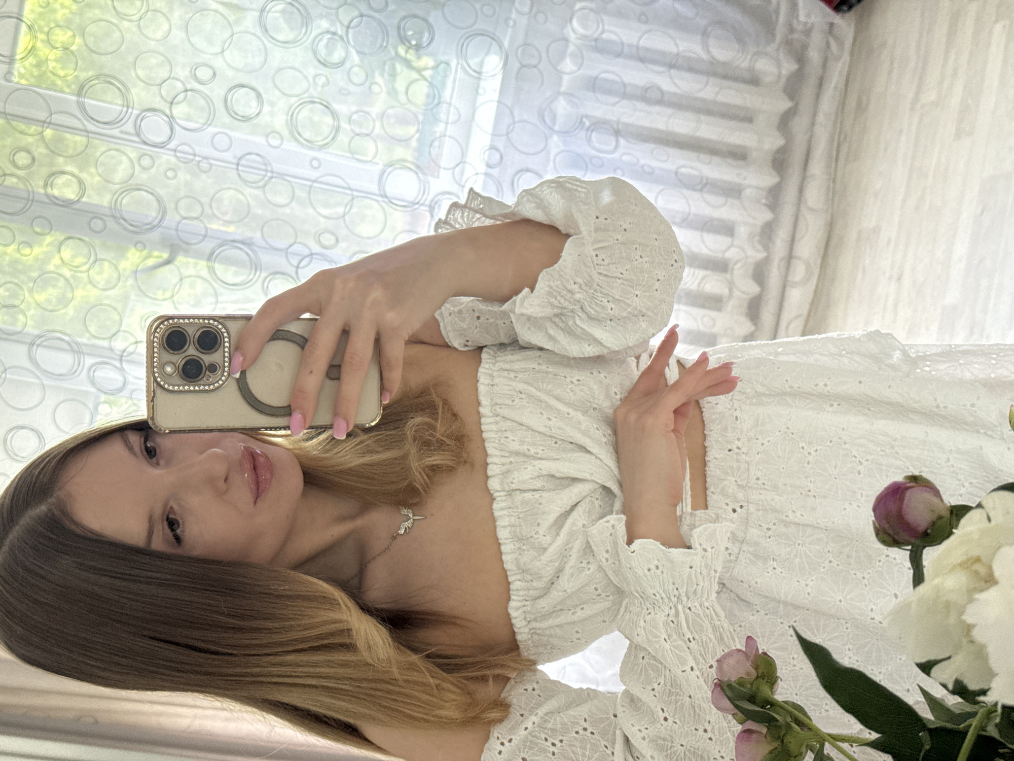 A young woman with long, light brown ombre hair is taking a selfie in a room with white circular patterned curtains. She is wearing a white off-the-shoulder top with ruffled sleeves and a delicate necklace with a winged pendant. Her left hand is holding a smartphone with a diamond-encrusted case, and her right hand is resting on her stomach. In the foreground, to the left, are several pink and white peony flowers. The room appears to be a domestic setting, possibly a bedroom or living room, with light filtering in through the window. The image does not contain any specific elements that would allow for an identification of the city of Октябрьское, Russia.
