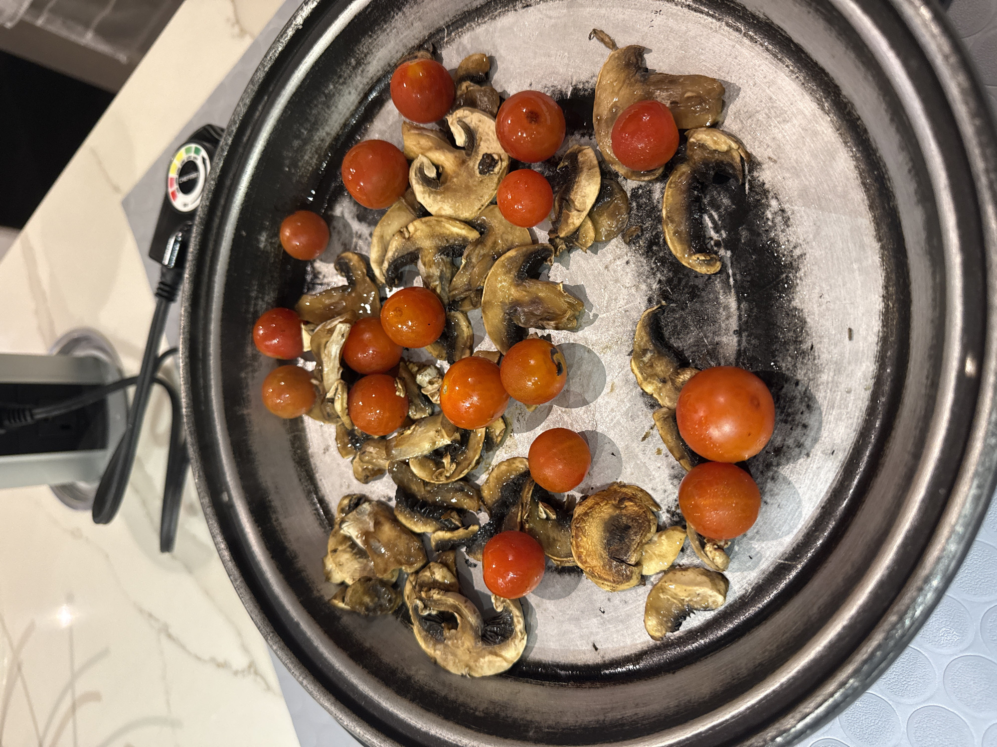 A top-down shot depicts a circular metal pan filled with sliced mushrooms and cherry tomatoes, some of which appear browned or slightly burnt. The pan is positioned on a light-colored surface, possibly a countertop, with a temperature control dial and associated electrical cord visible to the left. The setting appears to be indoors, likely a kitchen. There is no information to suggest this image was taken in San Bernardino Tlaxcalancingo, Mexico.