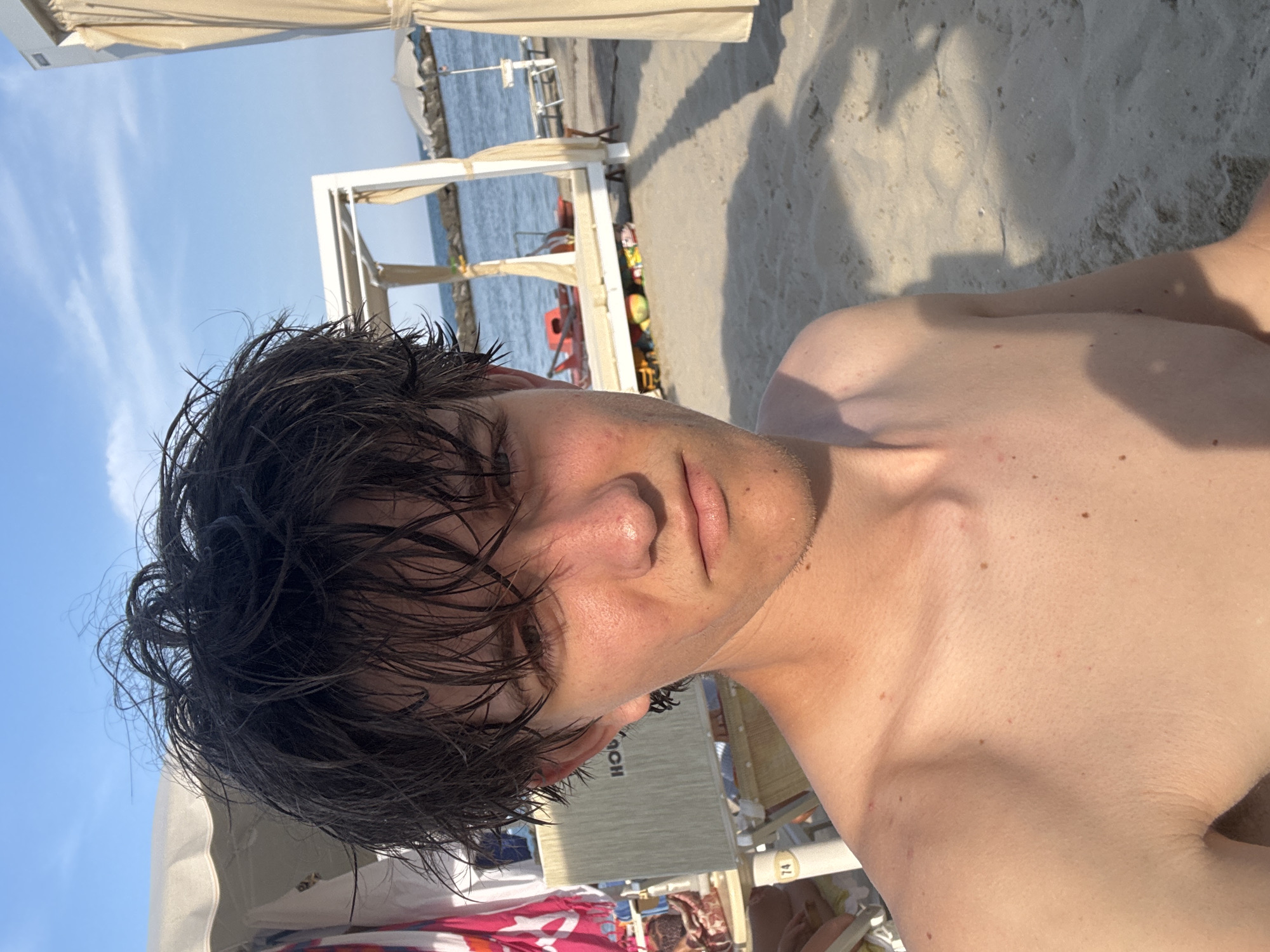 The image shows a young person lying on a beach. The person has dark, wet hair and is looking towards the camera with a relaxed expression. They are shirtless, and their skin appears sun-kissed.
The background reveals a beach setting with sand and the sea. There are lounge chairs and umbrellas, suggesting a sunny day at a resort or beach establishment. The water is visible, with some ripples on its surface. Shadows cast by umbrellas and structures indicate it is likely daytime, possibly late afternoon given the warm light.
Some text is visible on a white pole in the background, which reads "74". Other text, "HOCH," is partially visible on a light-colored surface behind the person. The setting appears to be in Cupra Marittima, Italy, as indicated by the location context. The overall mood is one of leisure and enjoyment of a summer day at the beach.