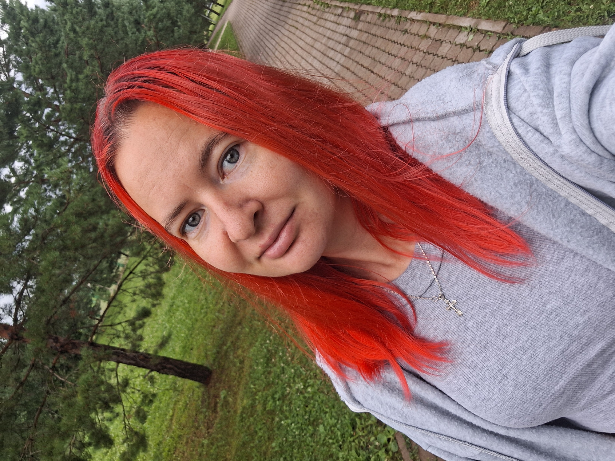 A mid-shot, outdoor, eye-level, selfie is taken from the chest up of a fair-skinned woman with bright red hair. She is wearing a grey t-shirt and a grey zippered hooded sweatshirt, and a silver cross necklace. Her light blue eyes are looking forward. The background consists of a paved walkway lined with grass and trees. The location is the city of Atamanovka, Russia.