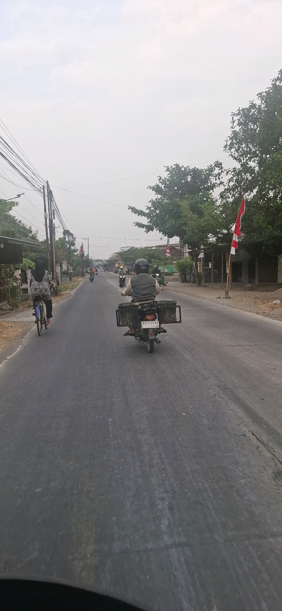 The image captures a view of a two-lane asphalt road in Desa Gagaksipat, Indonesia, under a hazy sky. A person on a bicycle, wearing a patterned headscarf and top, is in the left lane, moving away from the camera. In the right lane, also moving away, a person on a motorcycle wears a helmet and a gray vest over dark clothing. The motorcycle is equipped with two large, dark rectangular boxes attached to the rear, one on each side of the wheel. The license plate of the motorcycle is visible as "AD-2568-LK". Further down the road, several other motorcycles are in motion. The roadside features vegetation, including trees and bushes, and on the right side, a building with a red and white flag is visible, possibly the Indonesian flag. Utility poles with numerous wires are present along the left side of the road, and some smaller buildings and houses line both sides of the street. The overall atmosphere suggests a daytime scene with some atmospheric haze.