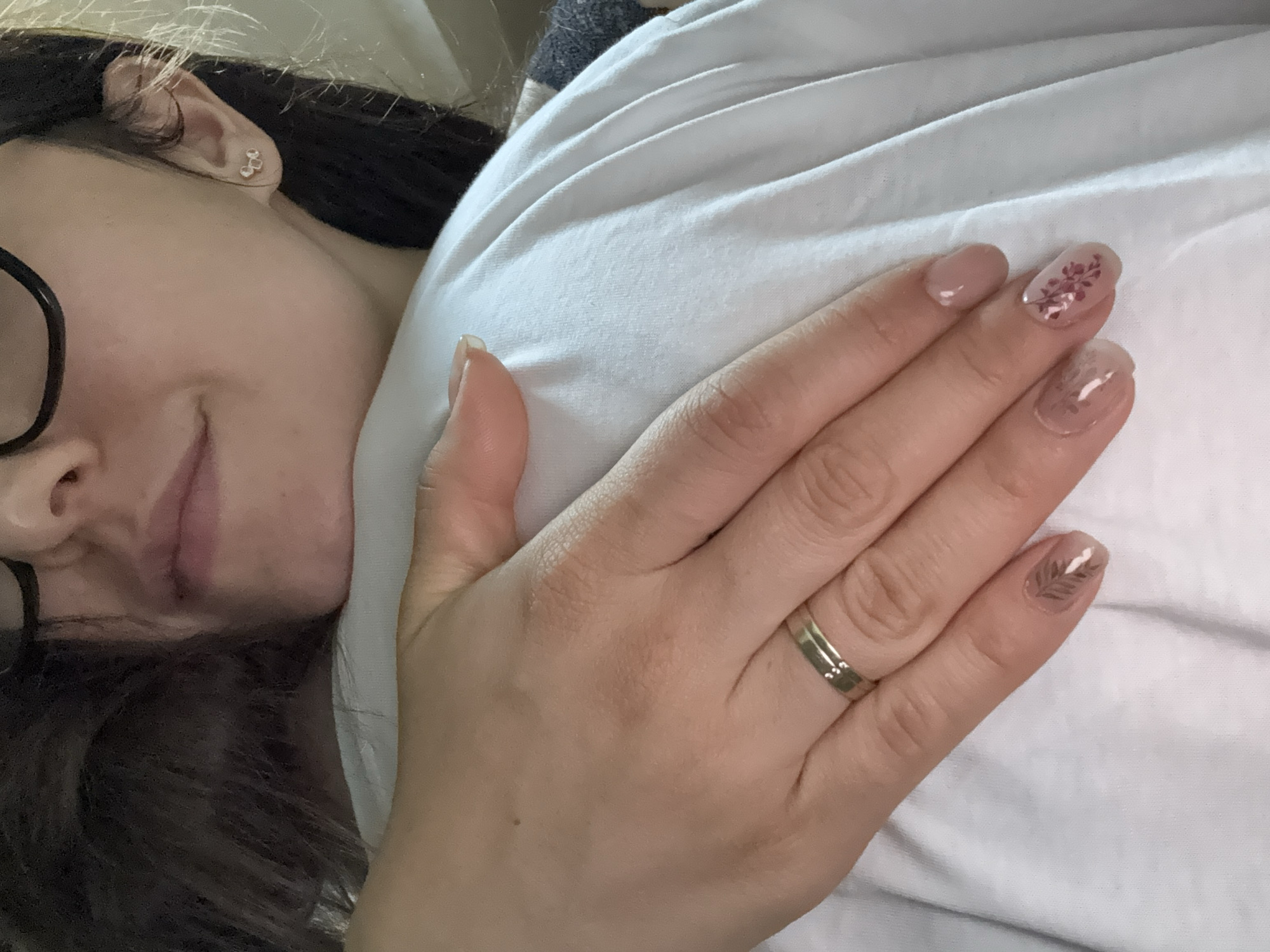 A person lies on a white surface, possibly a bed, with dark hair and glasses partially visible on the left side of the image. The person's face is slightly blurred. The right side of the image features a hand resting on the white surface, adorned with a gold ring on the ring finger. The fingernails are decorated with floral and leaf designs. The location in the city of Troyan, Bulgaria, cannot be determined from this image.