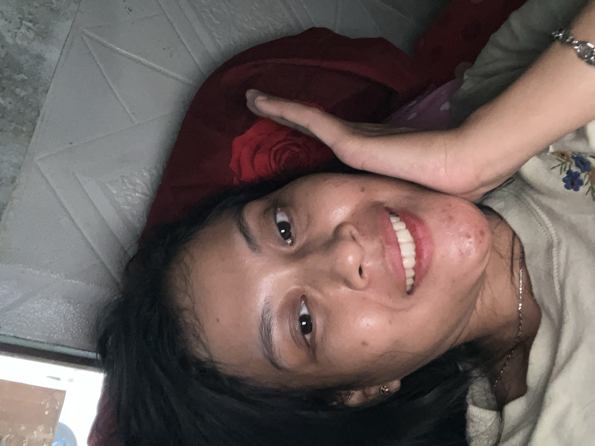 The image is a close-up selfie of a young woman lying down. She has dark hair and is smiling, with her right hand resting on her cheek. She is wearing a light-colored t-shirt with a floral design. Behind her, a red pillow with a rose pattern is visible, and a textured gray surface appears in the background. There is no information in the image to determine the location in Kecamatan Pati, Indonesia.