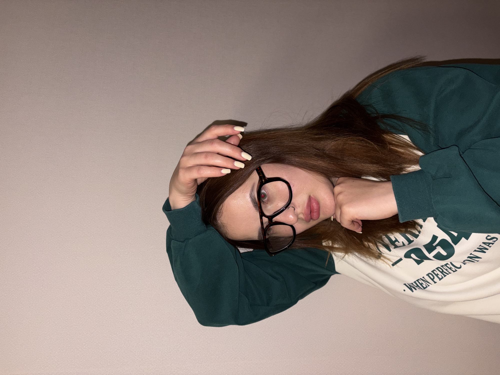 The image shows a young woman with long, reddish-brown hair, wearing thick-framed, dark tortoiseshell eyeglasses. She is positioned against a plain, light beige wall, looking directly at the camera. She wears a two-toned sweatshirt, featuring dark green sleeves and a primarily off-white body. Her left arm is bent with her hand near her head, displaying light yellow nail polish on her fingers, while her right hand is gently placed near her chin. A small stud piercing is visible on her left nostril. Her expression is neutral to slightly pensive, with a subtle pout. Artificial lighting illuminates the scene, suggesting an indoor setting, possibly during the evening. Partially visible text on her sweatshirt includes "-95" and "WHEN PERFECTION WAS".