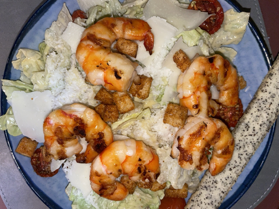 A blue plate holds a shrimp Caesar salad. Five large grilled shrimp curl amidst romaine lettuce, croutons, and parmesan shavings. Two sun-dried tomatoes add a touch of color. The lettuce is coated in a creamy Caesar dressing. Beside the salad is a breadstick covered in poppyseeds. The presentation suggests a tasty and savory meal.