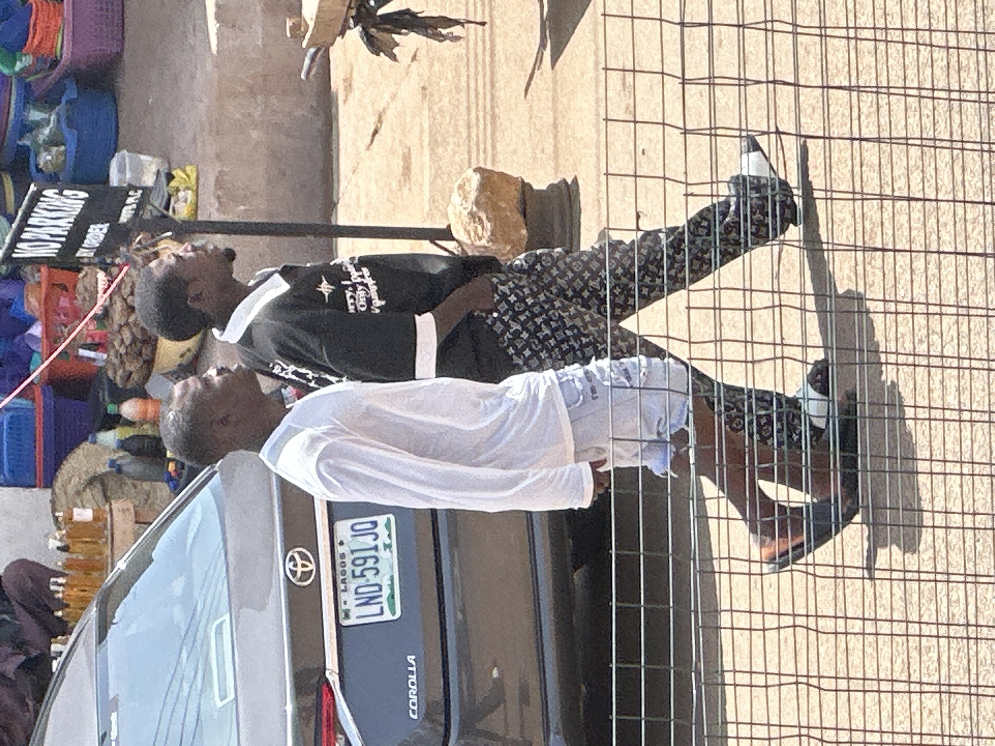 Two Nigerian men walk past a grey Toyota Corolla in Bukuru, Nigeria. The man on the left wears a white long-sleeved shirt and light blue denim shorts. The man on the right wears a black long-sleeved shirt with a white collar and a black and white patterned fabric. Both men are wearing dark-colored footwear. In the background, a "NO PARKING" sign is visible. Various items are displayed on tables, suggesting a market or roadside vending area. The men are walking behind a wire fence.