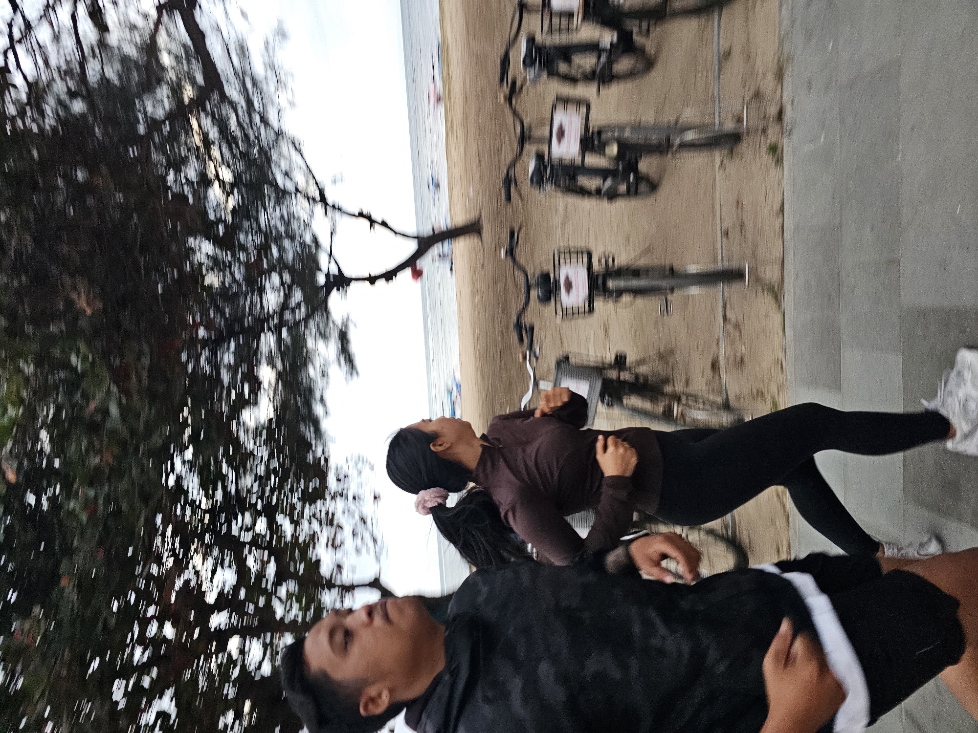 In Denpasar, Indonesia, two individuals, a man and a woman, are captured in motion, running along a paved walkway adjacent to a sandy beach. The man, positioned on the left, is wearing a dark, patterned hooded jacket and black shorts with a white trim. The woman, to his right, is in athletic wear, a dark long-sleeved top and black leggings, with her hair tied back in a ponytail. Behind them, several bicycles are parked in a row on the sandy beach, some with baskets. The background reveals the sea and a hazy horizon, suggesting an outdoor, possibly coastal, environment. The overall impression is one of dynamic movement, with motion blur indicating the fast pace of the runners.