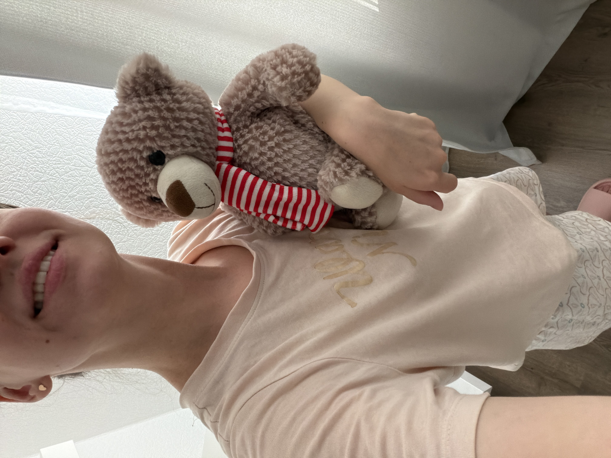 A medium shot shows a person holding a stuffed teddy bear. The person is wearing a light pink t-shirt with gold lettering and white patterned shorts. The teddy bear is brown with a red and white striped scarf. The person's face is visible, smiling. The background consists of white curtains and a wooden floor. The image was taken indoors.