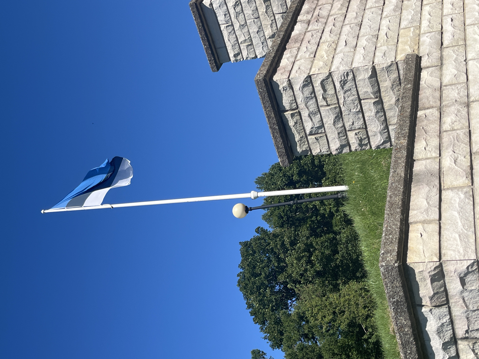 An Estonian flag flies on a white flagpole against a clear blue sky. The flag is composed of three horizontal bands of equal width: the top band is blue, the middle band is black, and the bottom band is white. The flag is positioned on the right side of the image, with its tail end curving to the left.
In the foreground, a light-colored stone wall with rough-hewn blocks forms a raised platform. The wall has a weathered appearance with grey capping stones along its top edges. Behind the wall, a patch of green grass slopes upwards.
To the left of the flagpole, a black lamppost with a round, cream-colored light fixture is visible, positioned on the grassy slope. Behind the lamppost and the grassy area, a dense canopy of green trees forms a backdrop.
The image appears to have been taken on a bright, sunny day. The overall setting suggests an outdoor public space, possibly a park or memorial in Toila, Estonia, given the presence of the Estonian flag. There are no people visible in the image.