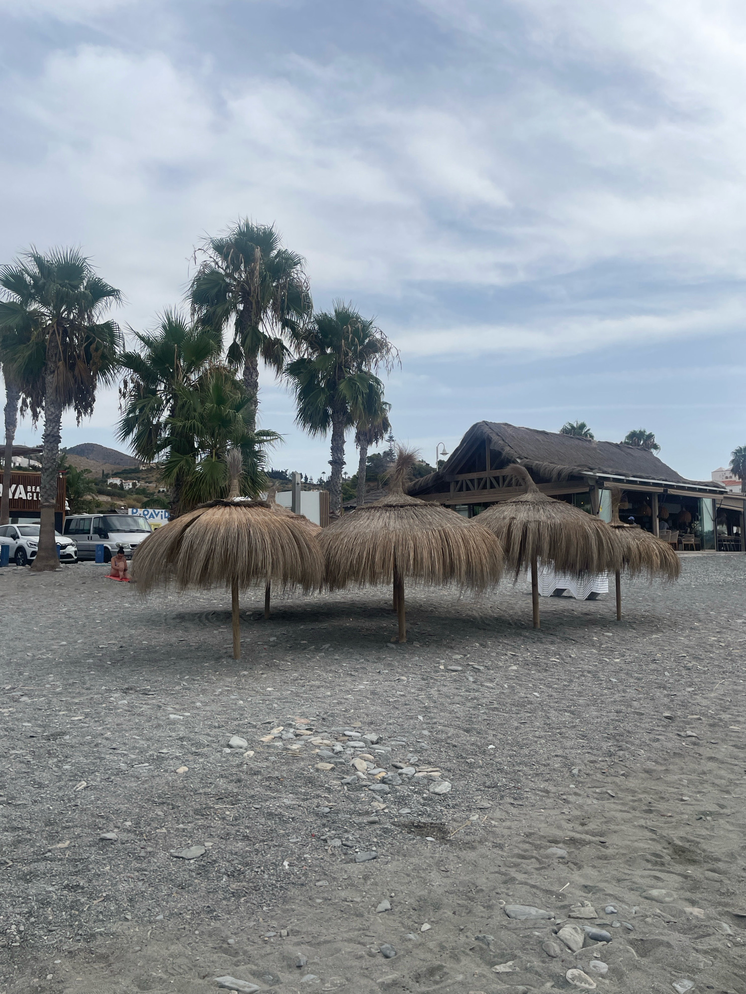 The image depicts a pebble beach in La Herradura, Spain, under a cloudy sky. In the foreground, several thatched umbrellas stand on wooden poles, casting shadows on the gravelly surface. A few birds are visible near the umbrellas. Behind the umbrellas, a beachside restaurant with a thatched roof and large glass windows is situated. Palm trees are scattered across the scene, with more visible in the background, along with some hills. Several cars and a van are parked on the road behind the beach, and a person is sitting on the beach. The overall atmosphere is serene and typical of a coastal resort.