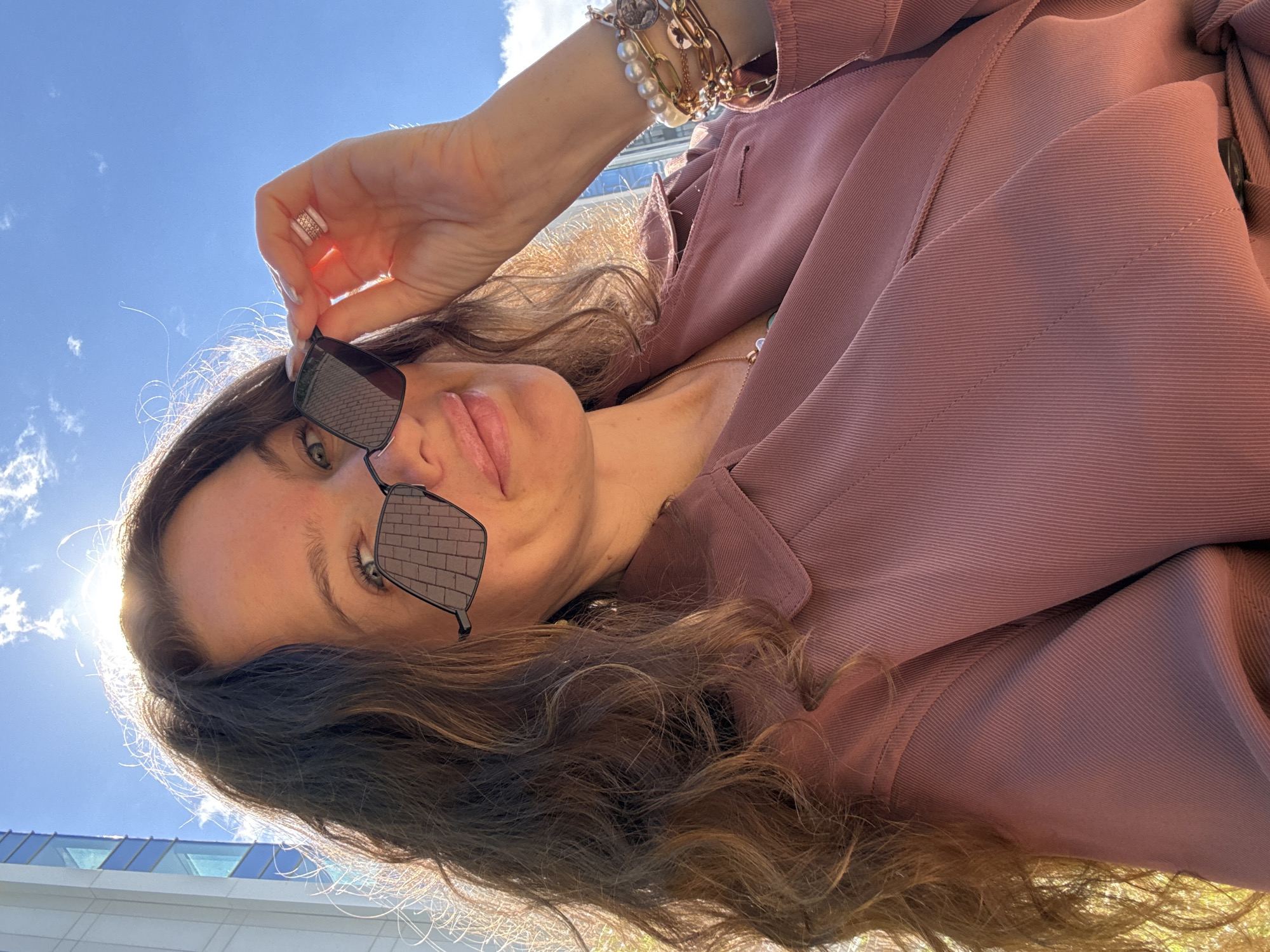 A young woman with long, wavy brown hair is taking a selfie outdoors. She is wearing sunglasses with a geometric frame, a pink jacket, and several bracelets on her right wrist. The sun is shining brightly behind her, creating a lens flare effect. In the background, a modern building and a clear blue sky with some clouds are visible. The woman is smiling and holding her sunglasses with her hand. The scene appears to be in a city, but there is no specific indication of the location being Moscow, Russia.
