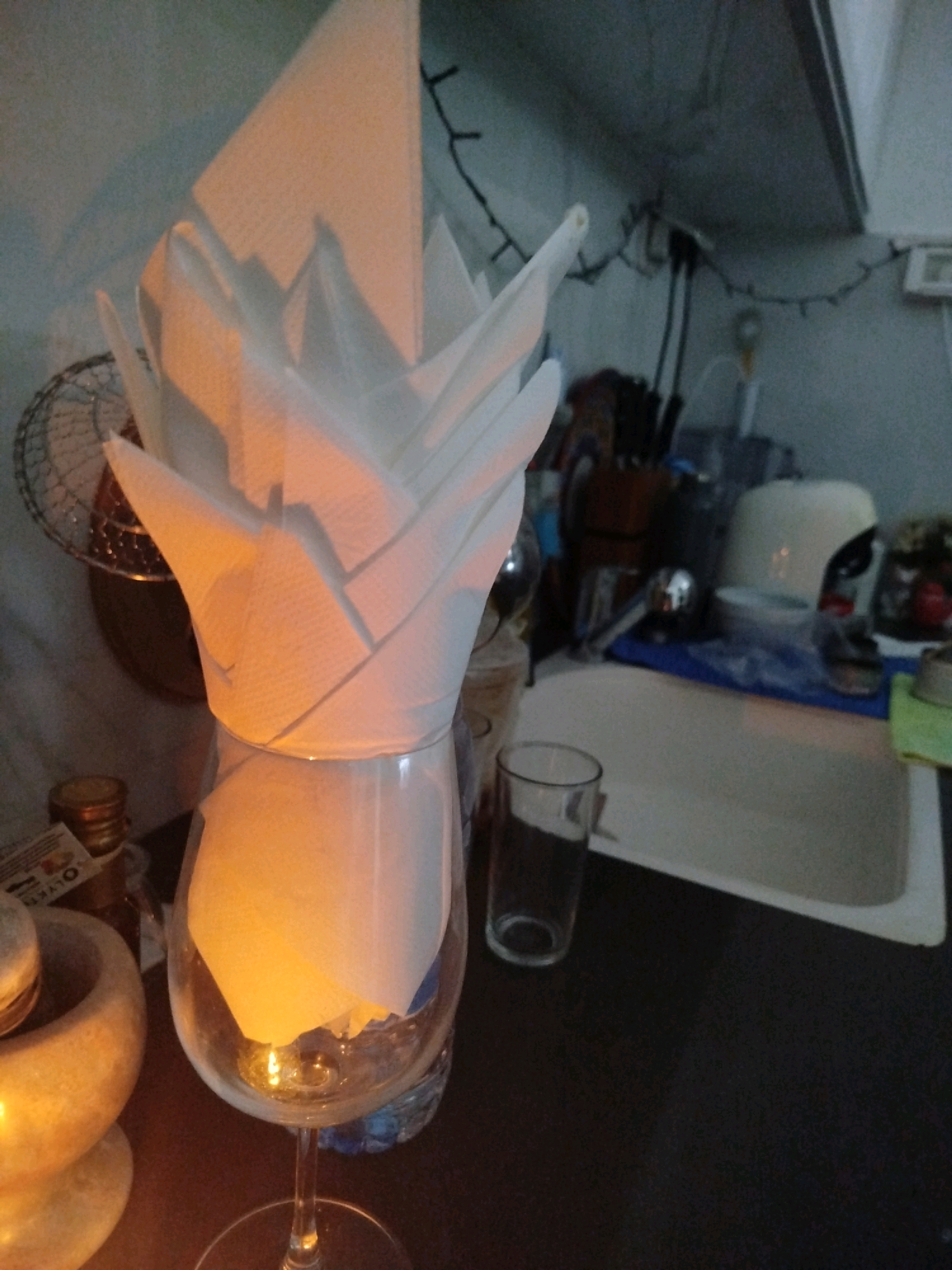 In a dimly lit domestic environment, possibly a kitchen or dining area, a white paper napkin is artfully folded and placed inside a wine glass. The napkin is folded into a multi-layered, petal-like structure, catching the warm light. The wine glass, which is clear and rests on a stem, contains the napkin and appears to be illuminated from within or by ambient light, casting a soft glow.
To the left of the wine glass, a light-colored mortar and pestle are visible, suggesting a preparation area. Behind the wine glass, a wire rack is partially seen, and further back, a sink area with various items, including a faucet and what appears to be a countertop, is present. The background also shows some string lights, adding to the ambiance. The overall scene is one of careful arrangement and a subtle play of light and shadow.
This image is not identifiable as being located in Ashkelon, Israel. There are no specific geographical markers or contextual clues to place it in that city.