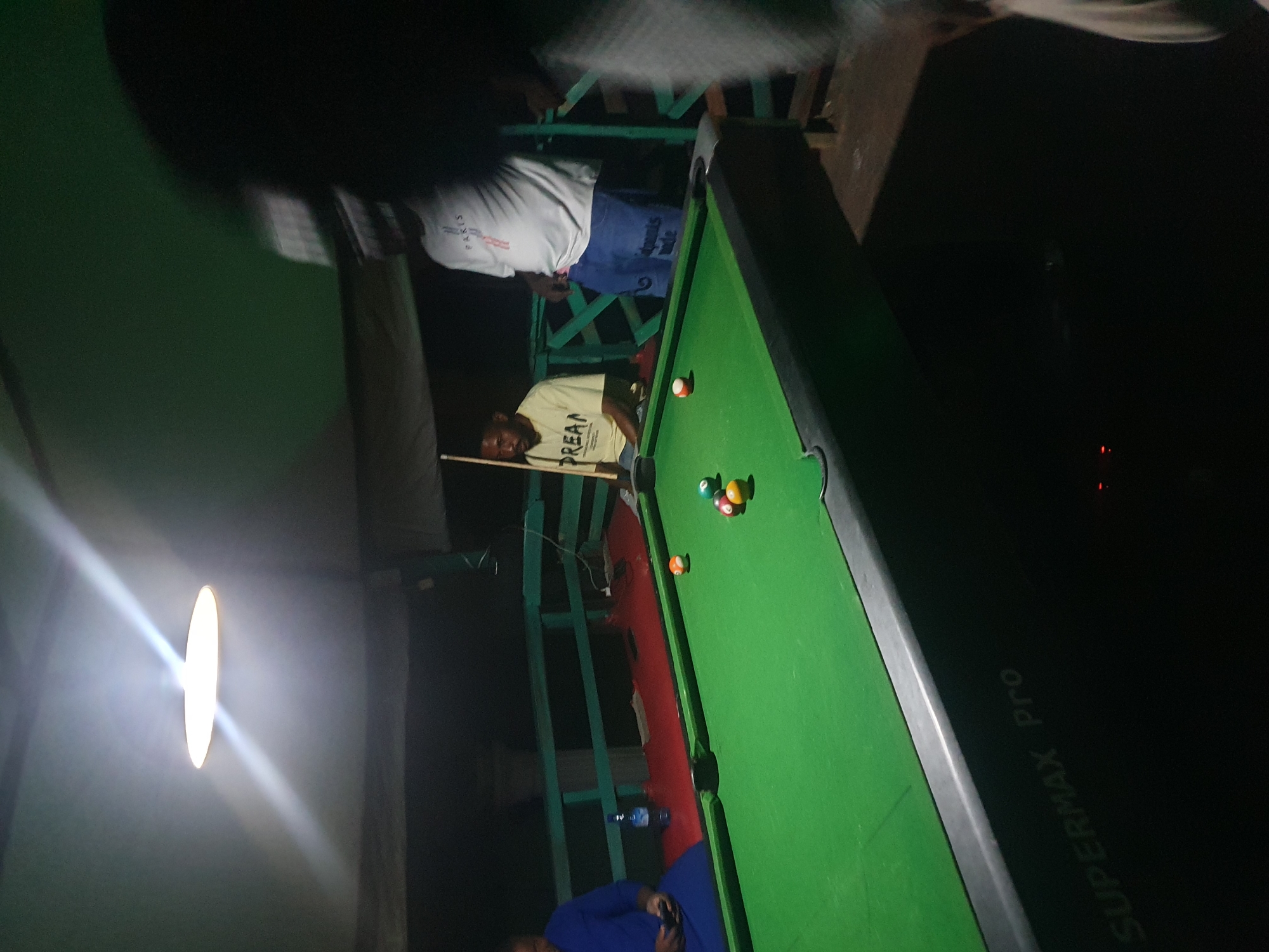 The image depicts an outdoor pool hall, likely in Lokoja, Nigeria, at night. A pool table with green felt and several colored balls is the central focus. Three individuals are visible: one person in a blue garment is seated to the left, appearing to use a mobile phone. Another person in a light-colored t-shirt with "DREAM" printed on it is seated behind the pool table, holding a cue stick. A third person, wearing a white t-shirt with "PARIS" printed on it and blue jeans, is standing. The environment appears to be somewhat rustic, with a green railing and a wooden structure visible in the background. A bright overhead light illuminates the scene. The overall atmosphere suggests a casual gathering for playing pool.