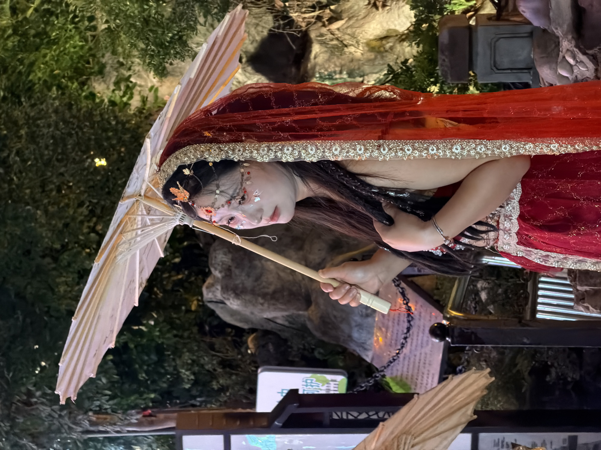 A young East Asian woman, dressed in a red and gold traditional costume with elaborate jewelry and makeup, holds an ornate white and tan umbrella. She is positioned outdoors at night, with a backdrop of foliage and rocks. To her left, a sign with Chinese characters is partially visible, suggesting a location within Yuzhong District, China. The scene appears to be part of a cultural event or performance.