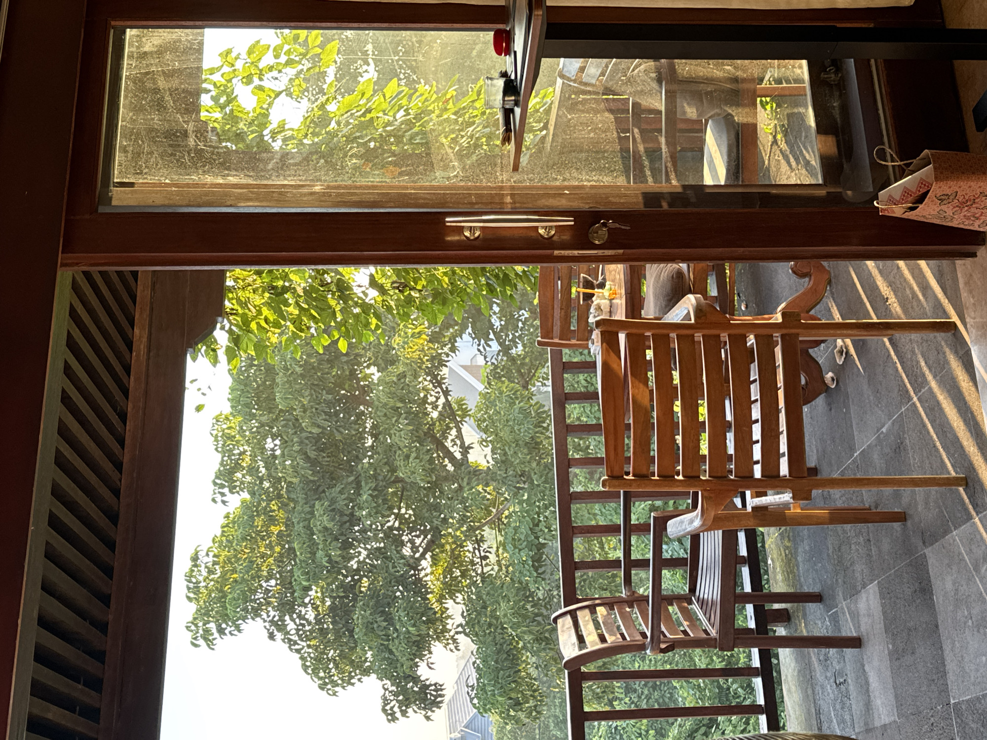 The photograph presents a view through an open doorway, likely in a building or establishment in Kuta Selatan, Indonesia. The doorway is framed by dark wood, with an upper window section that appears dusty or slightly fogged. Through this upper window, green foliage is visible against a bright, hazy sky, suggesting either early morning or late afternoon light.
Below the upper window, the doorway opens onto a space containing wooden furniture. A wooden chair is prominently featured, its slats and frame catching the sunlight. The space beyond the chair shows more greenery, possibly a garden or park area. The overall impression is one of an indoor-outdoor setting, with the doorway serving as a transition between a building's interior and the external natural environment. The presence of wood and foliage suggests a design aesthetic incorporating natural materials and a connection to nature. The floor surface is visible and appears to be made of gray tiles or stone.
