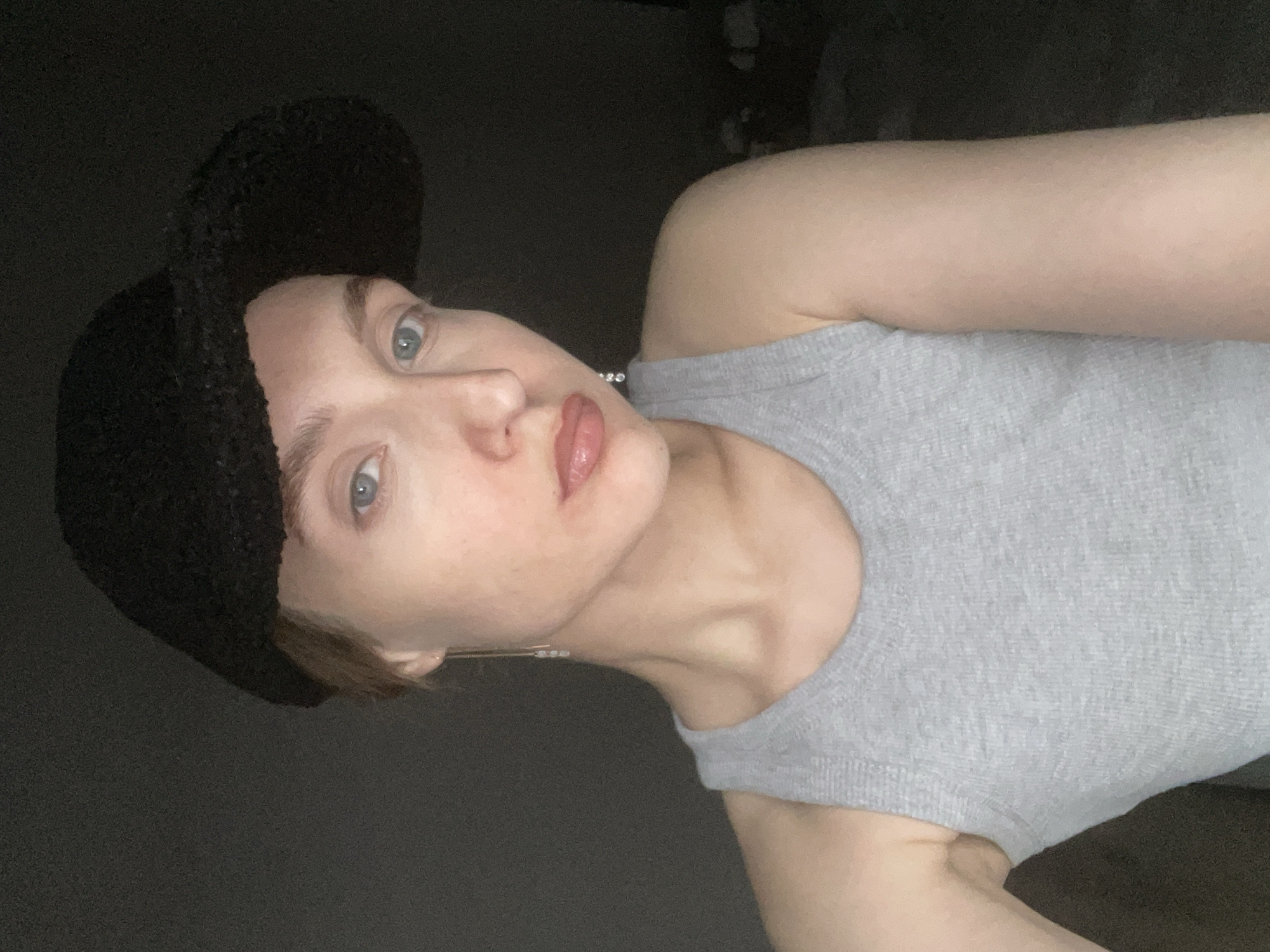 A woman with fair skin and blue eyes is wearing a black hat and a gray tank top. She is facing the camera with her head tilted slightly. The background is dark and indistinct. The location cannot be determined from the image.