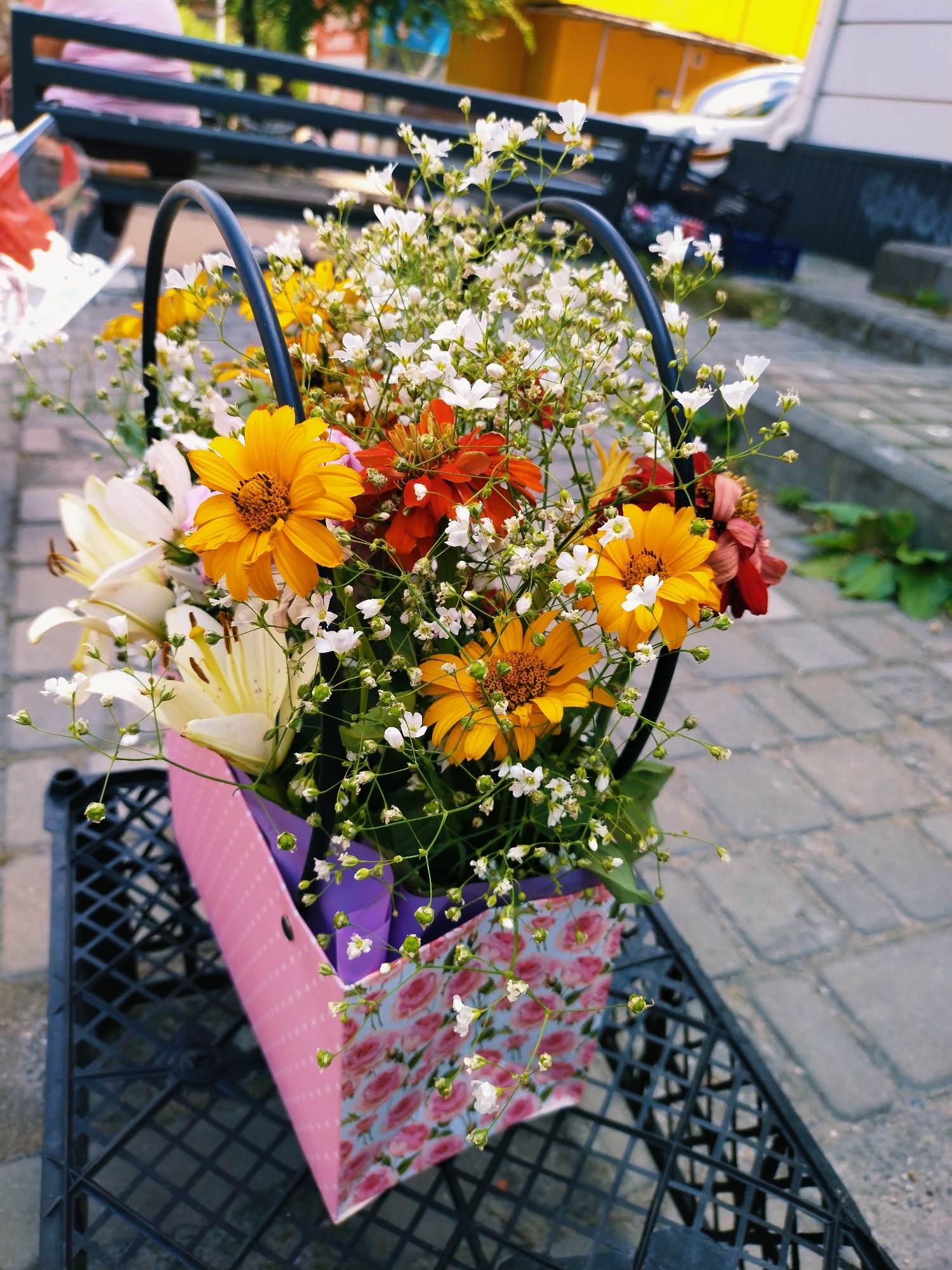 A bouquet of flowers is displayed in a decorative pink bag with a floral pattern. The bouquet is a mix of yellow daisies, white lilies, and small white flowers, with some red and orange zinnias also visible. The flowers are arranged in a black basket handle. The bag is placed on a black plastic crate, which sits on a paved surface made of grey interlocking bricks. In the background, a dark metal bench with a person in a pink shirt is visible, along with a yellow structure and some greenery. The scene appears to be outdoors, possibly in a public space like a park or a street, given the paving stones and bench. The lighting suggests it is daytime, with the sun casting some shadows. The overall mood is cheerful and vibrant, thanks to the colorful flowers. There is no visible text in the image.