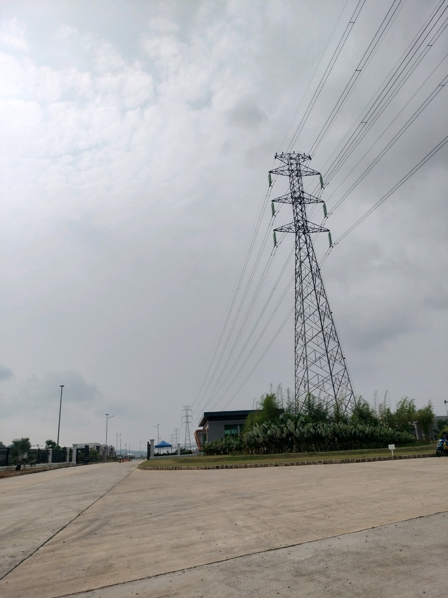 The image shows a power transmission tower in Batam, Indonesia, against a cloudy sky. The tower is tall and made of metal lattice structure, with multiple power lines extending from it. In the foreground, there is a paved road or an open area with concrete surface. Further back, there are some buildings and greenery, including bamboo plants. The scene appears to be an industrial or infrastructure area.