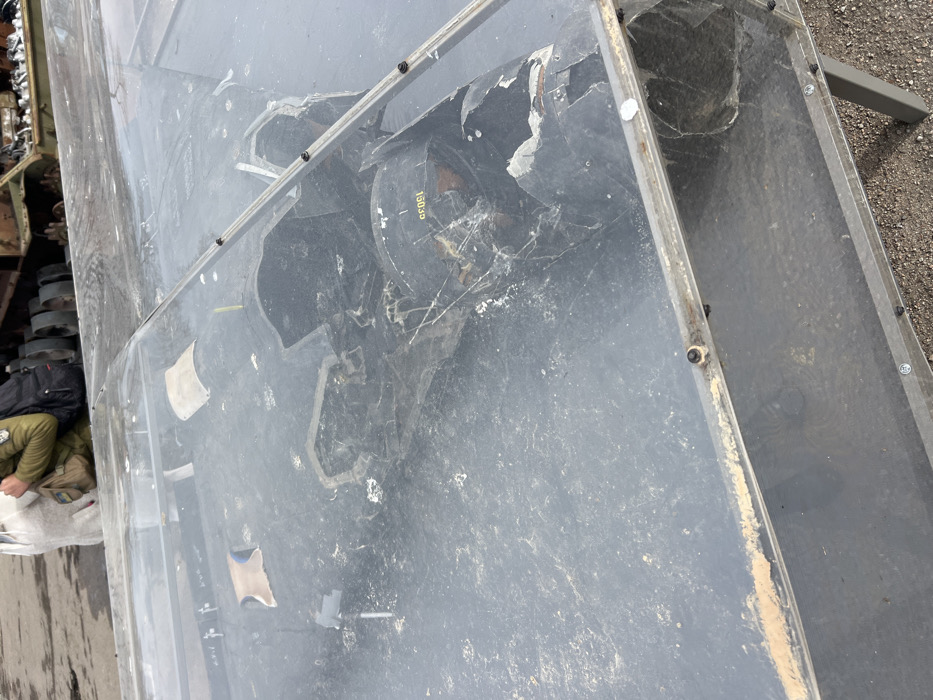 The image captures an outdoor exhibition in Odesa, Ukraine. The primary subject is a rectangular display case, constructed with a metal frame and clear, scratched plexiglass panels secured by bolts. Inside the case, resting on a dark, flat surface, are fragments of what appear to be a missile or rocket. A prominent, curved black piece of debris with yellow alphanumeric markings "1503G" is visible, surrounded by smaller, irregular metallic and composite shards and white particulate matter. In the upper-left background, two individuals are partially visible: one wearing a light-colored hoodie and holding a white bag, and another in a khaki jacket carrying a backpack and a tan-colored pouch. Behind these individuals, a large, dark, metallic structure consistent with military vehicle wreckage or industrial machinery is partially observed. The ground surface is a mixture of asphalt and gravel.