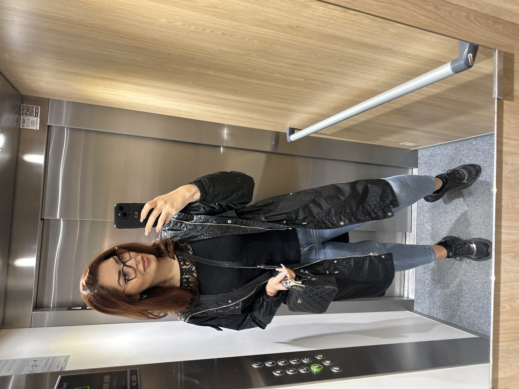 A woman is taking a selfie in an elevator. She is wearing glasses, a black top, light blue jeans, a black jacket, and black sneakers. She is holding a black smartphone and a black purse. The elevator has a wooden ceiling, stainless steel walls, a grey handrail, and a control panel with numbered buttons. A QR code and informational sticker are visible on the left wall. The location is possibly Sofia, Bulgaria, but this cannot be definitively confirmed from the image.