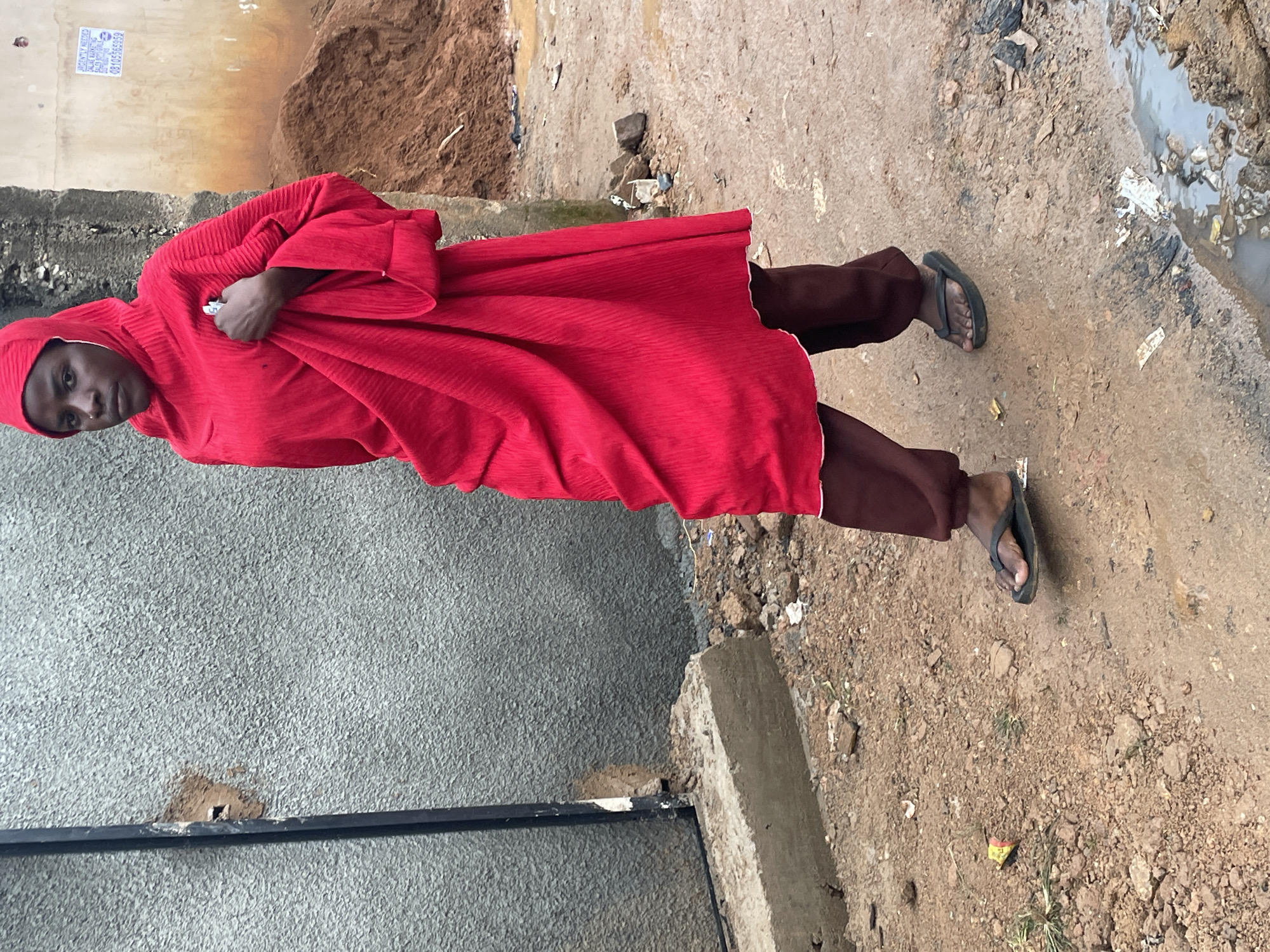 A young woman with dark skin, wearing a bright red hijab and a matching flowing dress over brown pants, walks across a muddy, unpaved street in Jos, Nigeria. Her dark flip-flops are visible on her feet as she navigates the uneven terrain, which is littered with debris and puddles of water. A rough, grey concrete wall with a dark vertical fixture is on her left. Behind her, a pile of reddish-brown dirt and a beige building with a faded sign are visible. The overall scene depicts a gritty, everyday environment in an urban setting.