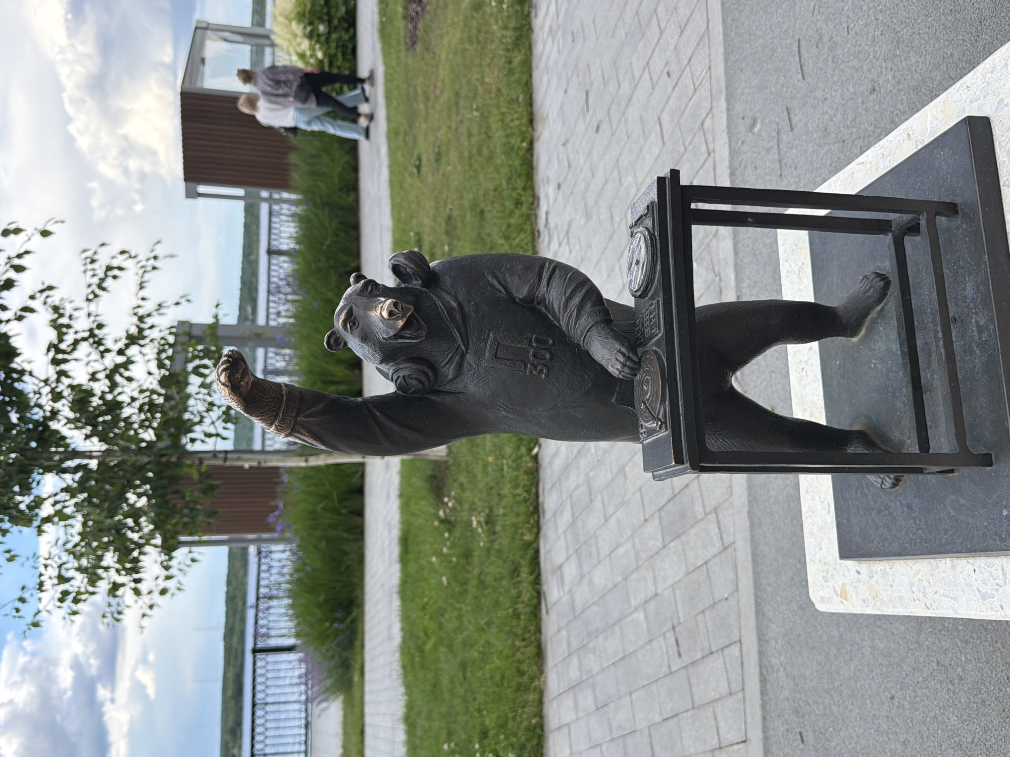 A bronze statue of a bear wearing headphones and a hoodie with the number "300" on it, stands behind a DJ setup. The bear is depicted with one arm raised in a fist. The statue is located in Perm, Russia, specifically on the embankment of the Kama River, near the Art Gallery. In the background, two women walk along the embankment, with a body of water and trees visible beyond. The ground is paved with gray tiles. The sky is partly cloudy.