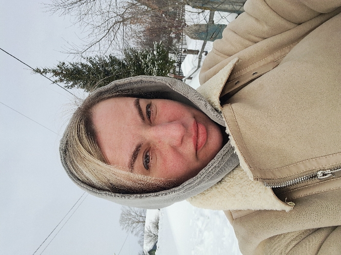 A female subject is depicted in a close-up outdoor selfie, situated in a snow-covered environment identified as the city of Полянки, Russia. She has fair skin with visible facial redness, and blonde hair partially visible beneath a light grey hooded garment. She is wearing a beige, shearling-lined winter jacket featuring a prominent metallic zipper, partially open. The subject looks directly into the camera with a neutral gaze. The background reveals an extensive layer of snow under an overcast sky. Visible elements include a snow-covered path, a dark green coniferous tree, bare deciduous trees, and a weathered wooden fence to the right. Utility lines traverse the upper portion of the sky.