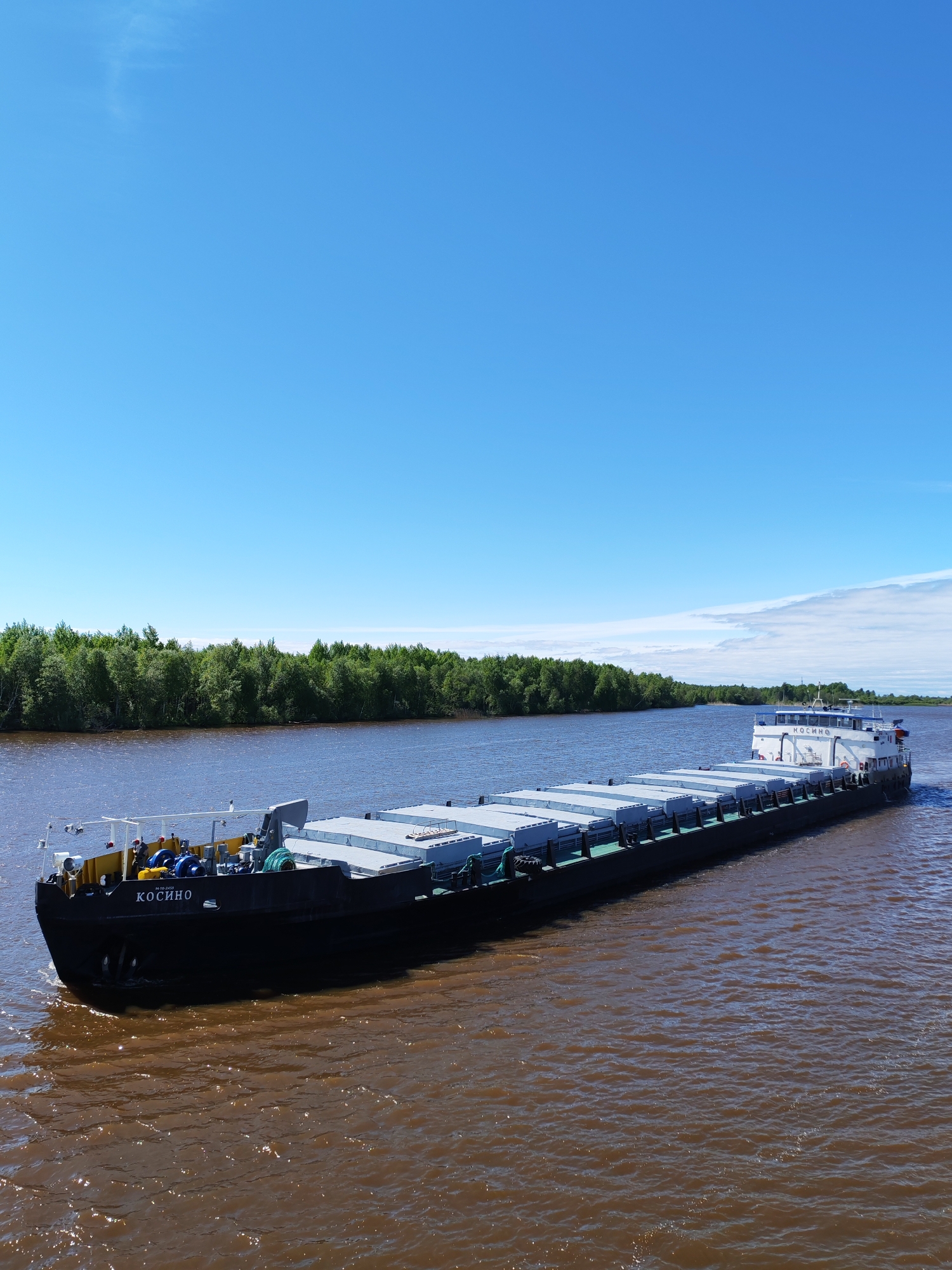 A barge, identifiable by the name "КОСИНО" (KOSINO) emblazoned on its bow, is seen navigating the river. The riverbanks are lined with dense green vegetation. The sky above is clear and blue. The location is Kargulino, Russia.