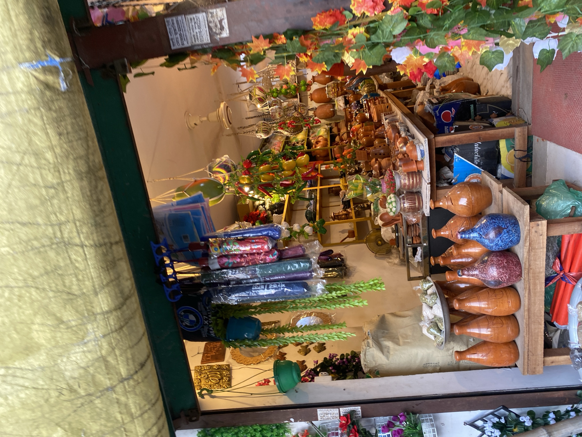 The image shows a roadside shop in Dhaka, Bangladesh. The shop is densely packed with a variety of goods. On the right, there is a display of earthen pots and jars of varying sizes and colors, arranged on a wooden rack. Further inside the shop, the shelving units are filled with pottery, decorative items, and other merchandise. The roof of the shop is adorned with artificial foliage and flowers. In the foreground on the left, there are stacked items wrapped in plastic, possibly textiles or similar goods. Outside the shop, there are floral decorations along the lower edge of the image.