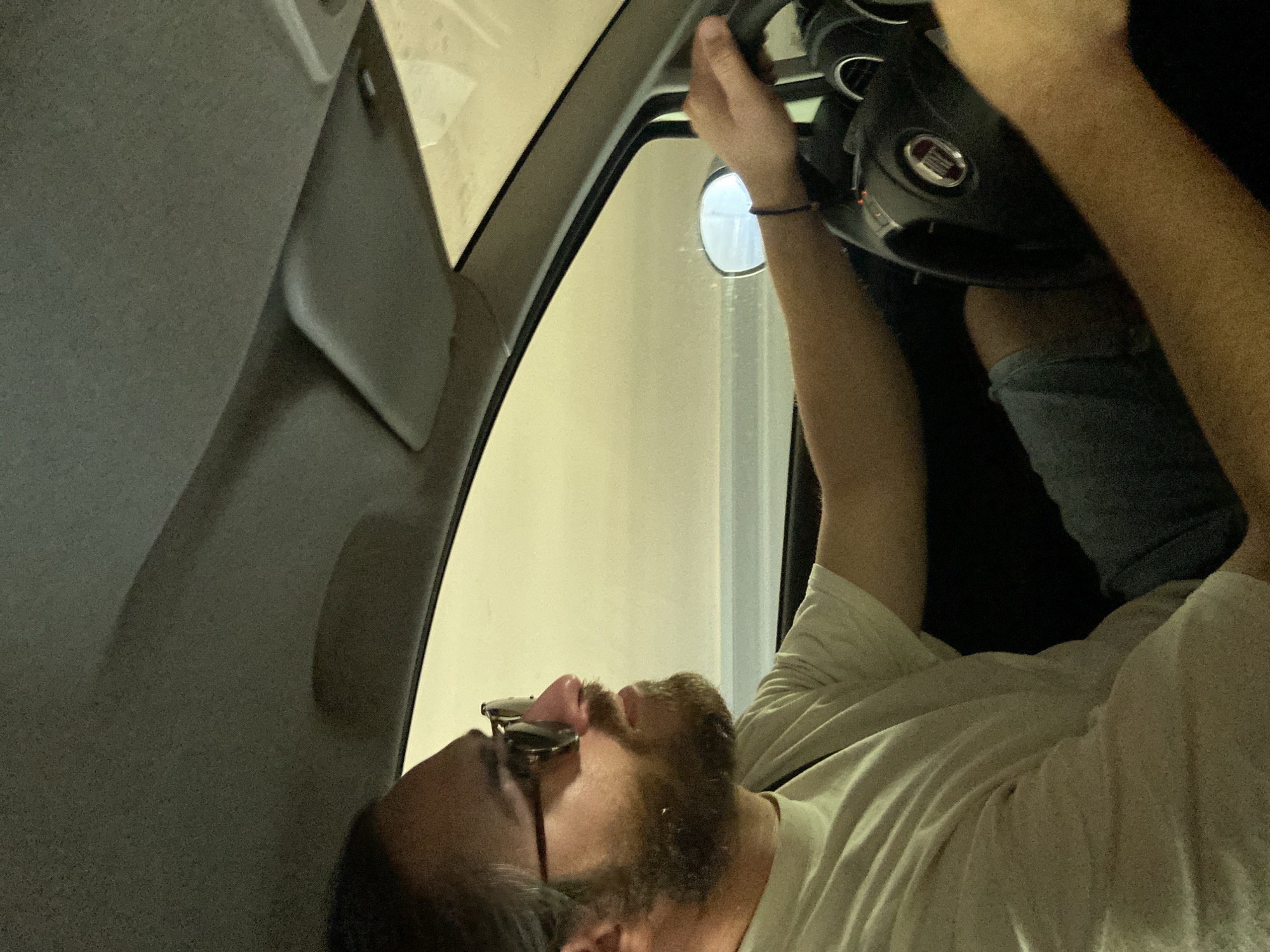 Inside a car, a bearded man wearing glasses and a light-colored t-shirt lies horizontally with his head near the front of the vehicle. He is holding onto the steering wheel with his hands. The rearview mirror is visible above him. The car interior is predominantly grey. The scene is likely set in Sotonići, Montenegro, based on location data.
